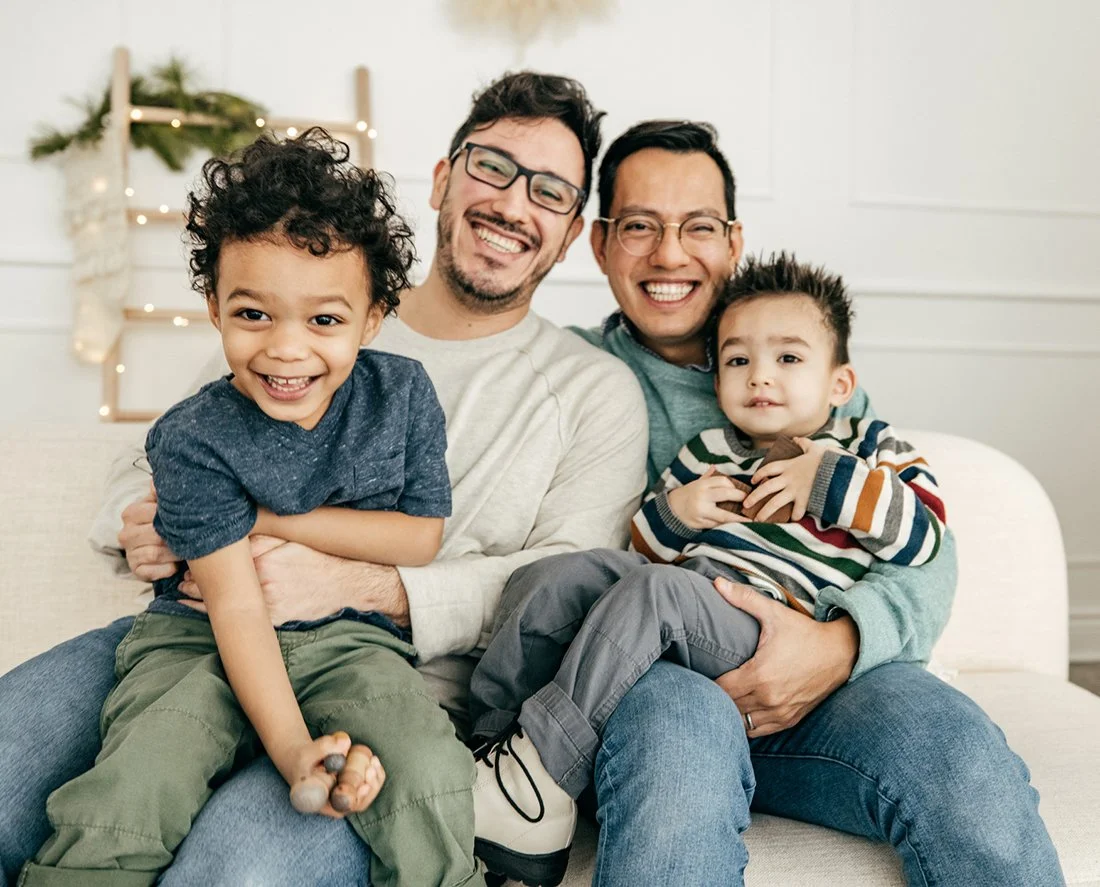 A young, mixed race gay couple sits on a beige couch with their two toddler sons