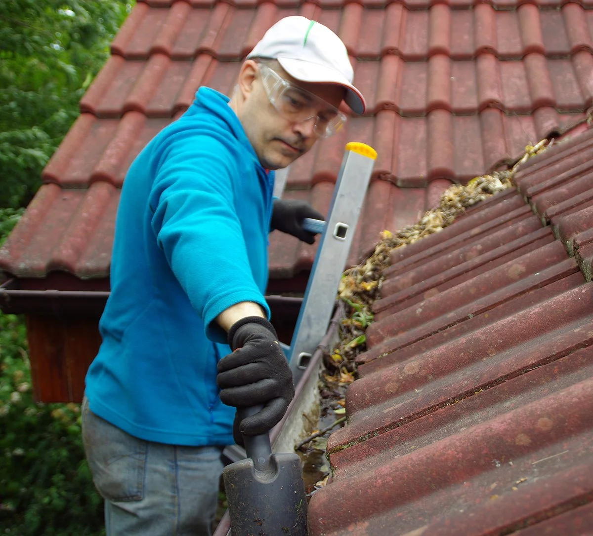 An older man wearing a blue sweater and white ballcap standing on a ladder cleaning the roof gutters