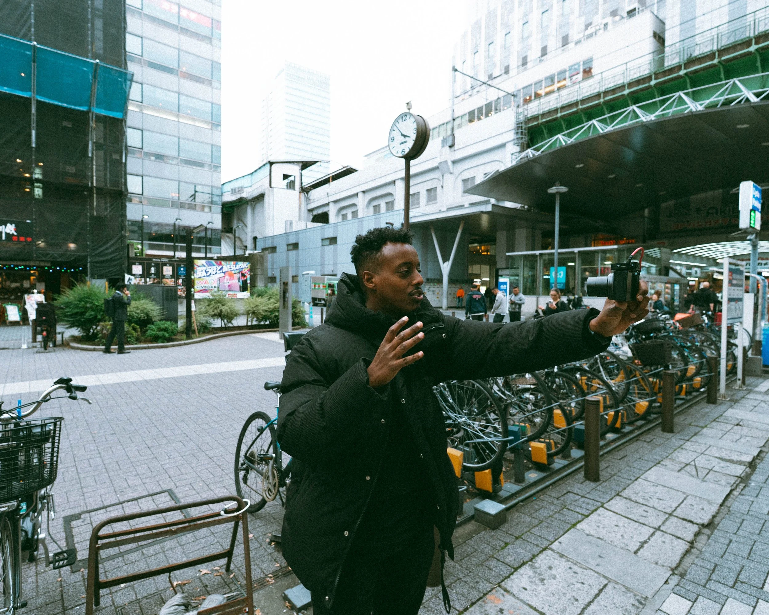 A man in a black jacket takes a selfie with a camera in an urban setting with bicycles and high-rise buildings.