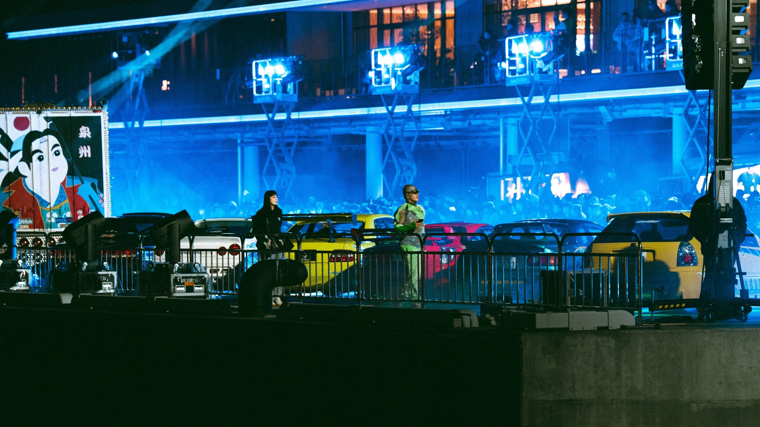 Nighttime scene at a parking lot with colorful cars, a woman and a person in a costume standing near the cars, and a crowd of people in the background illuminated by blue stage lights.