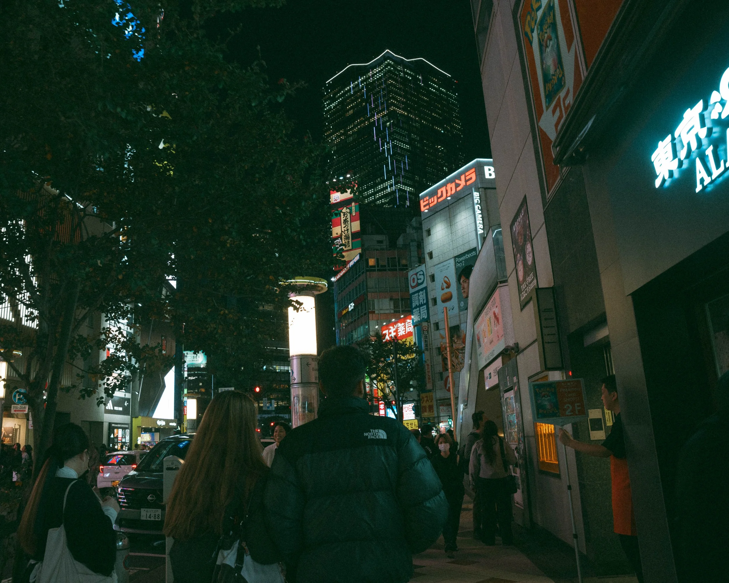 Nighttime city street scene with lit-up buildings, advertisements, and pedestrians walking, some wearing masks.