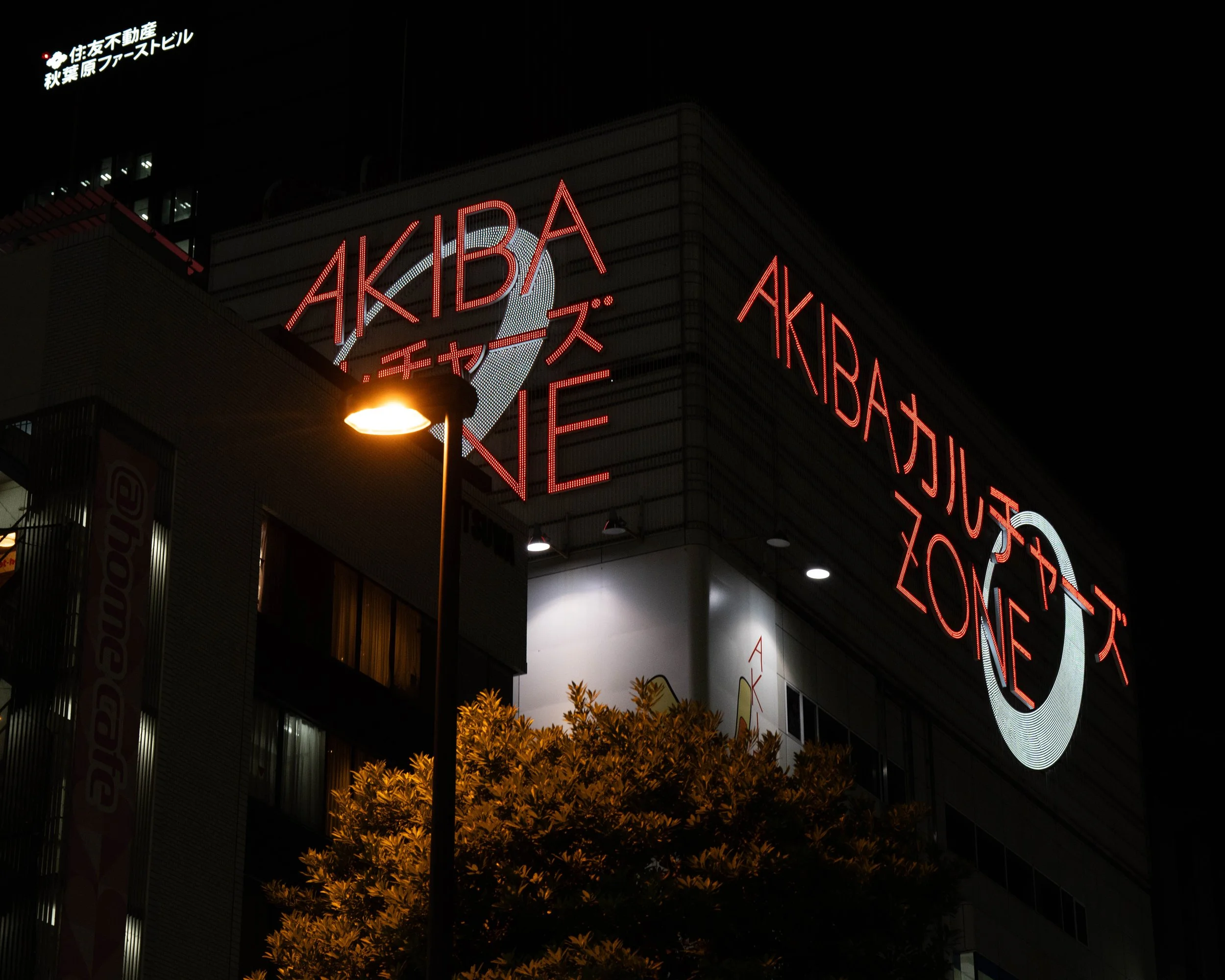 Night view of a building with illuminated red neon sign that reads 'AKIBA ZONE' in English and Japanese. A streetlamp and trees are visible in the foreground.