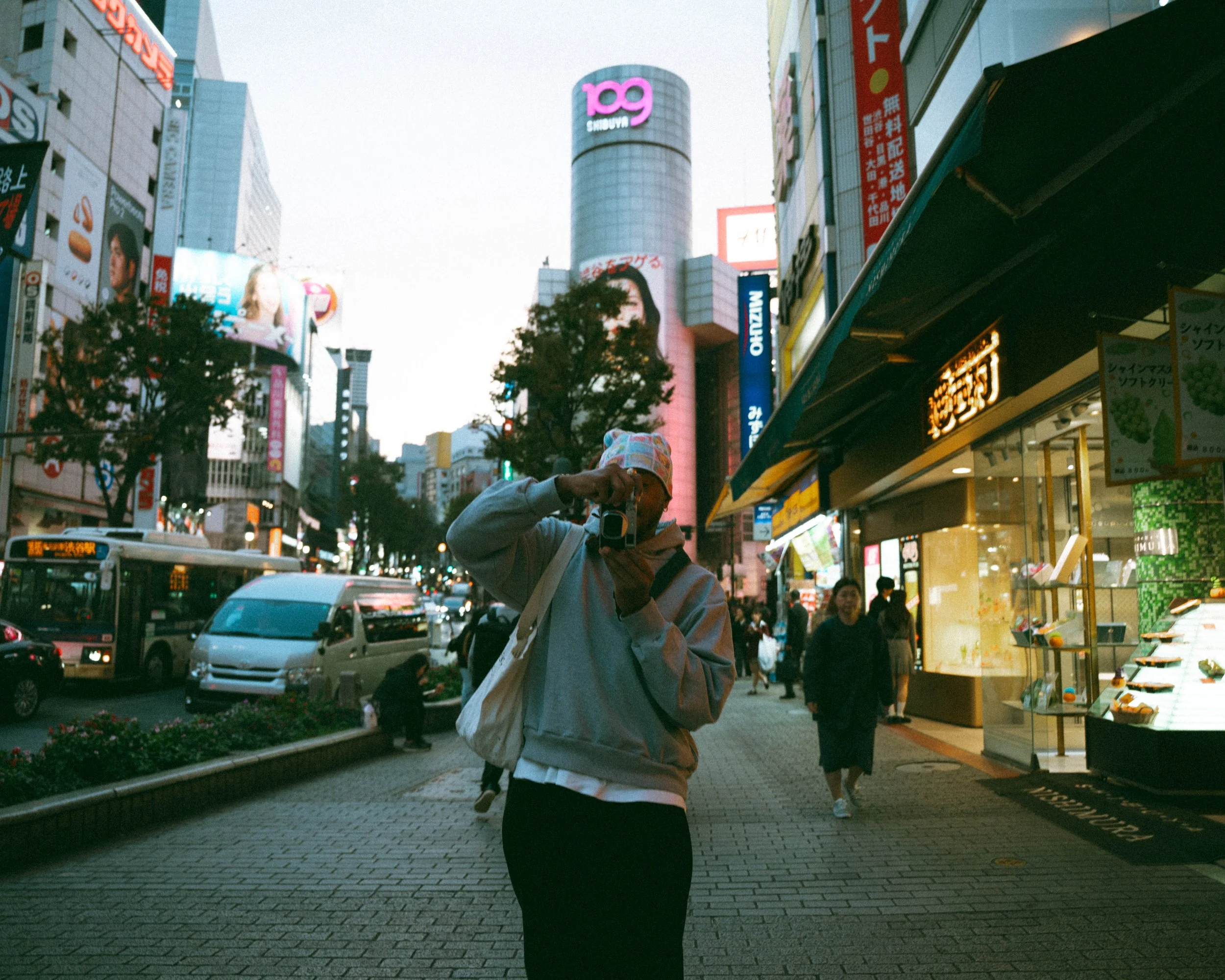 A person in a gray hoodie and colorful hat takes a picture with a camera on a busy city street at dusk, surrounded by illuminated storefronts, billboards, and pedestrians.