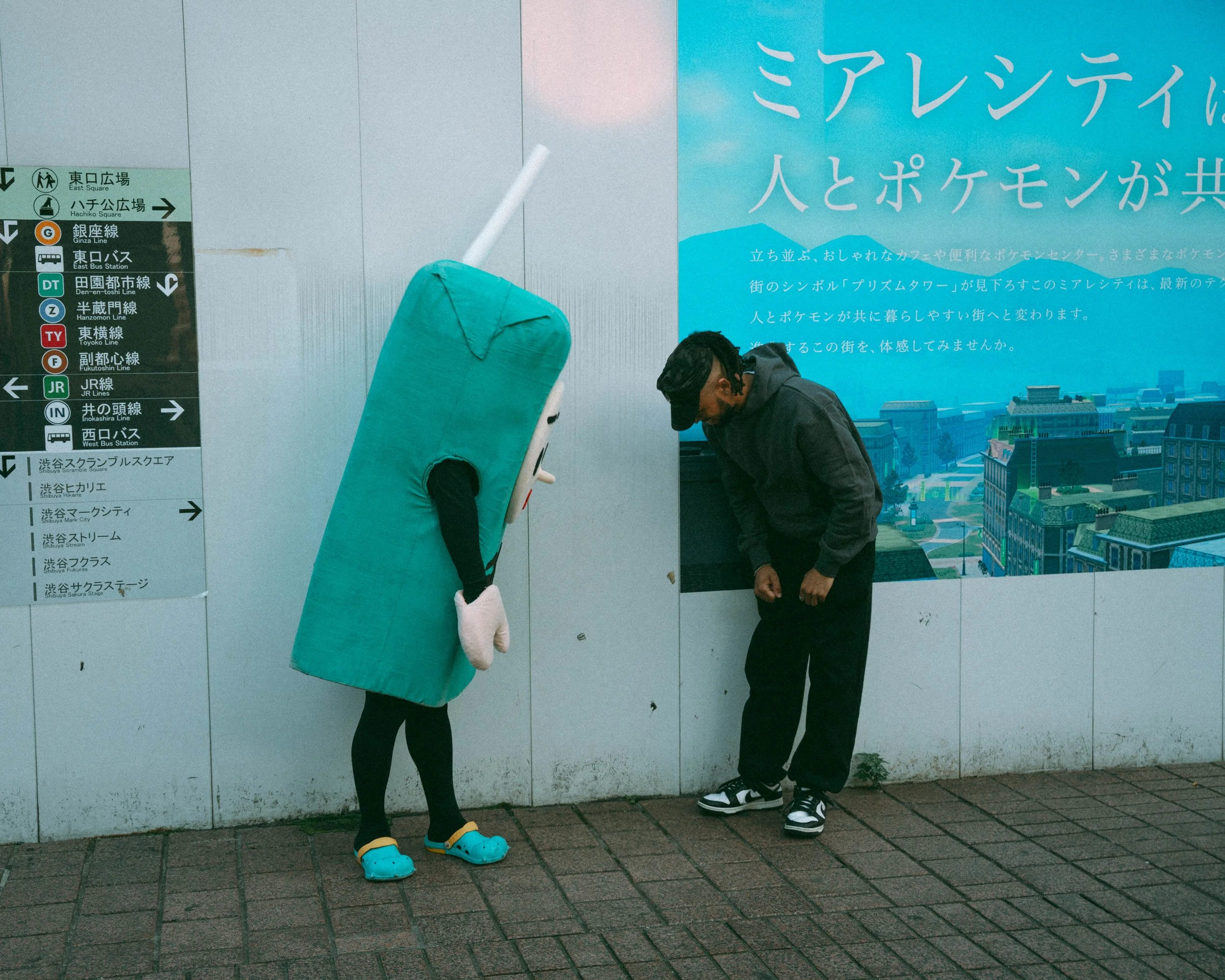 A person dressed in a camera costume talking to a man leaning forward in front of a wall with signs and a large blue poster with Japanese text and cityscape.