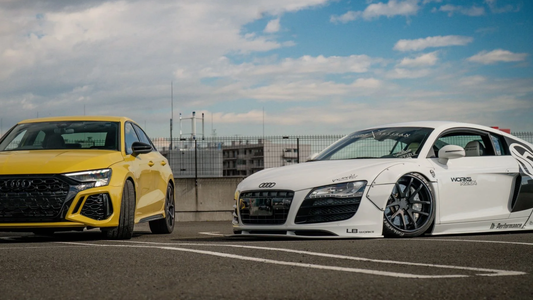 Yellow hatchback and white racing car parked in a lot under a partly cloudy sky.