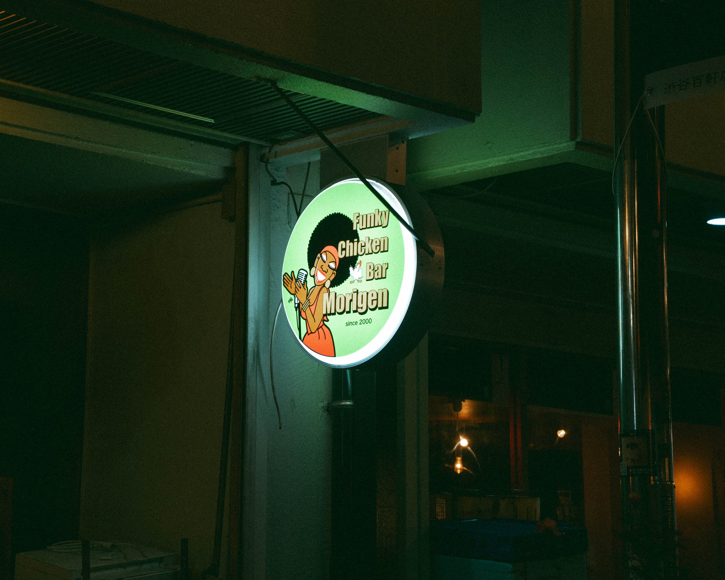 Illuminated sign reading 'Funky Chicken Bar Morigen' with a cartoon woman singing into a microphone and an Afro hairstyle, located outside at night.