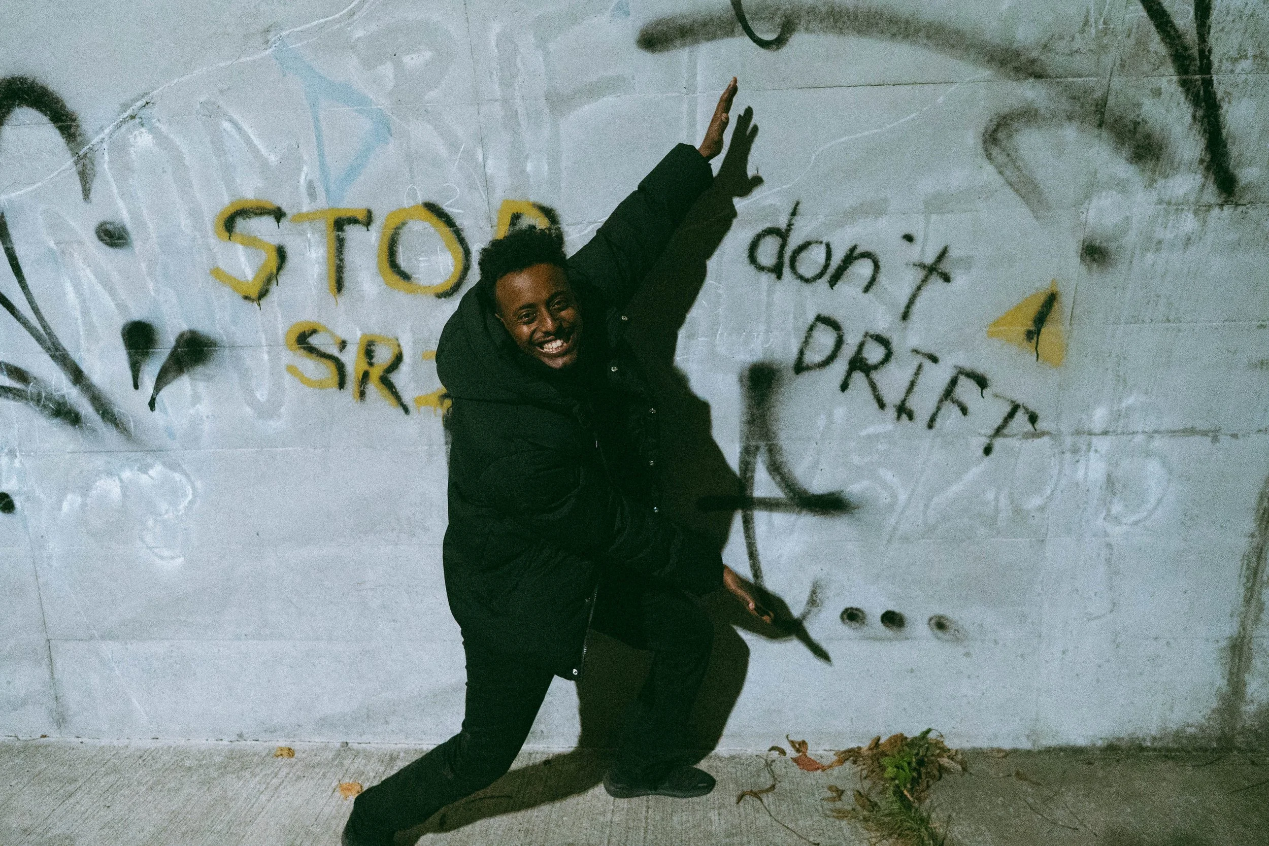 Person in black jacket smiling and pointing at a graffiti wall with the words 'STOP SR' and 'don't DRAFT' spray painted on it.