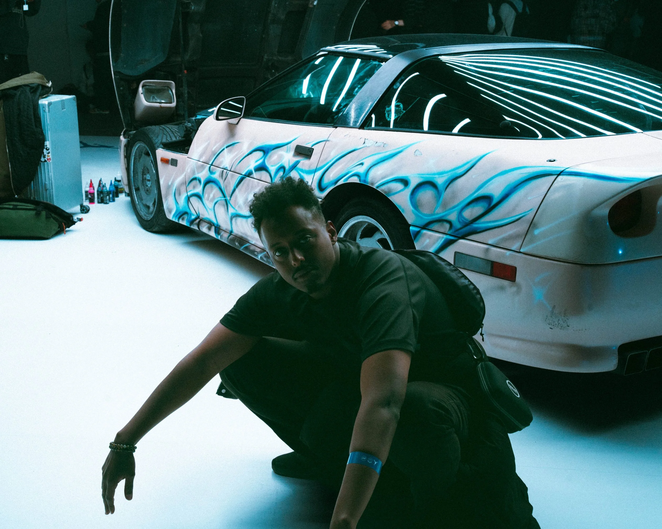 A person with curly hair crouching next to a white sports car with blue flame decals, in a dimly lit garage or event space with bottles and suitcase on the floor.