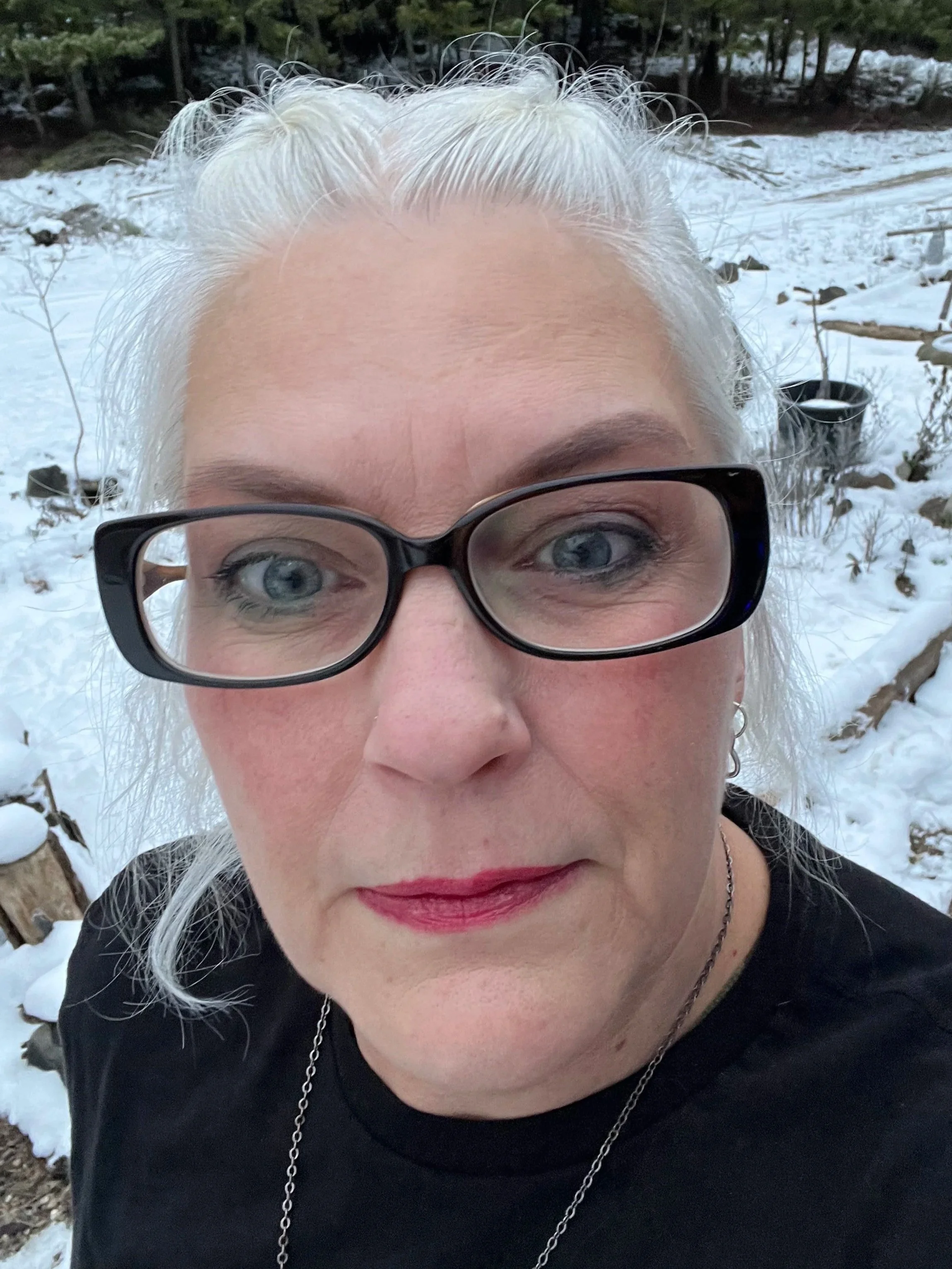 Close-up of a woman with white hair, glasses, and makeup, outdoors in a snowy landscape with trees and snow-covered ground.