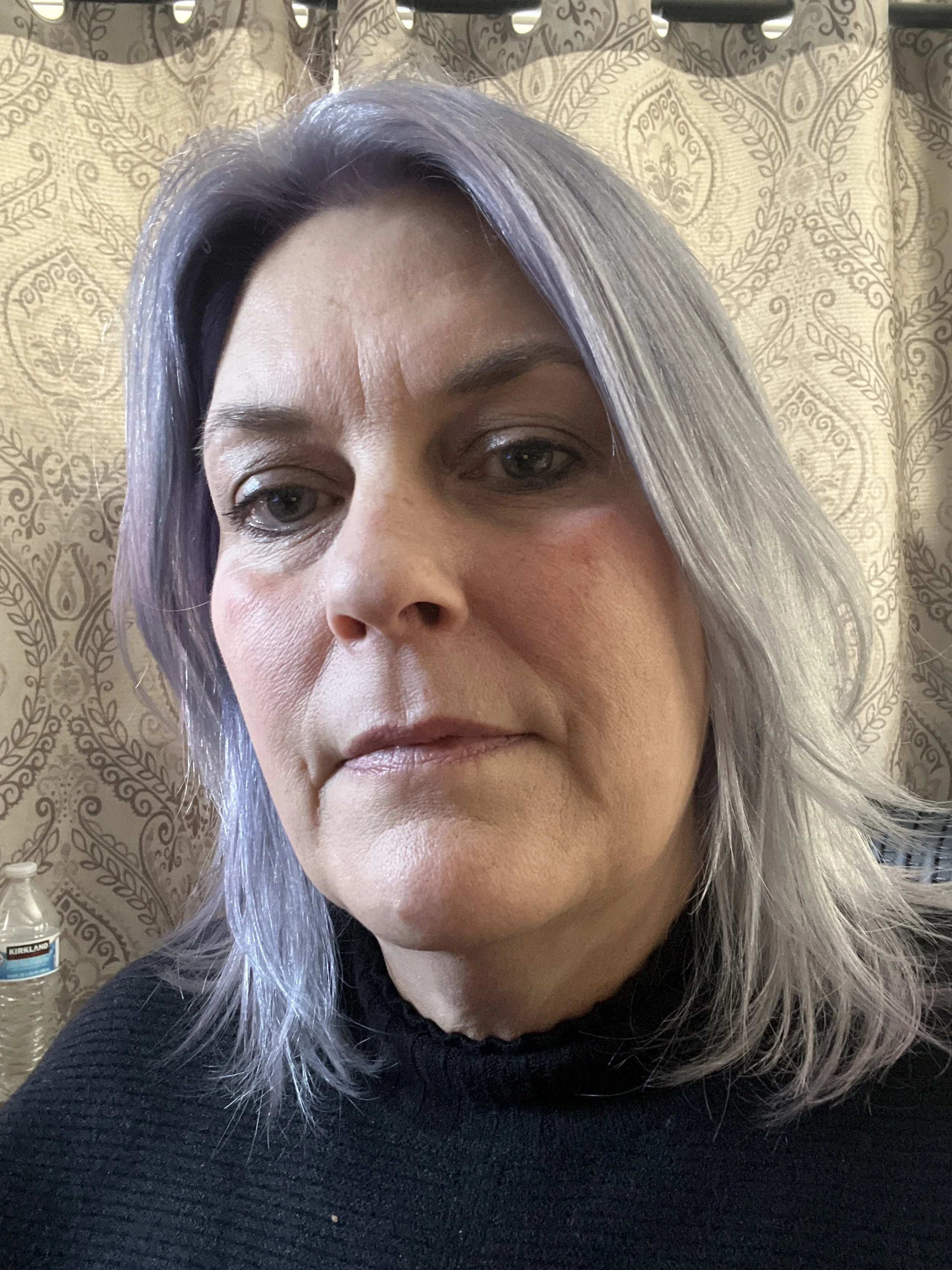 Close-up of a woman with shoulder-length grayish lavender hair and light skin, wearing a black top, in front of a patterned curtain.
