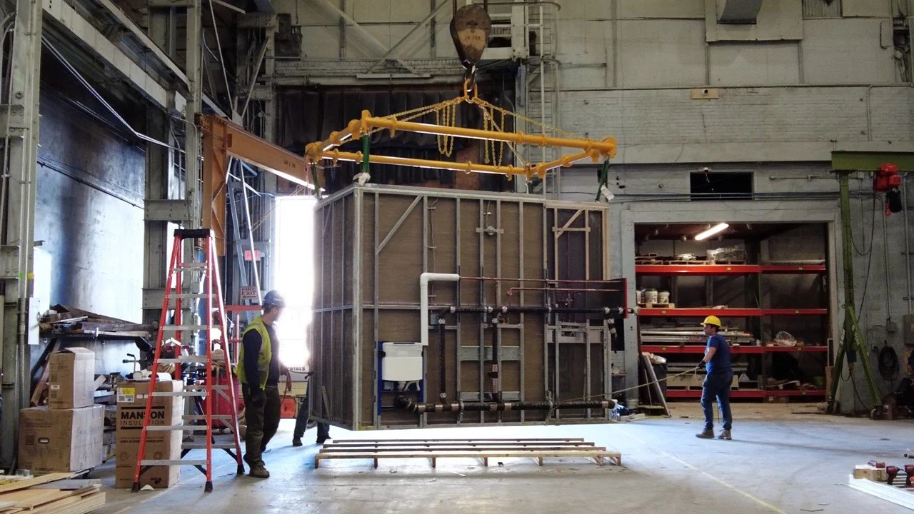 Workers are installing or inspecting a large industrial equipment or structure in a factory or warehouse, with one worker on the left near a red ladder and another on the right holding a measuring tape, while a large hoist or crane is lifting the structure in the center.