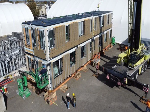A two-story modular building under construction on a flatbed trailer, with workers and a crane on site, surrounded by industrial equipment and storage domes.