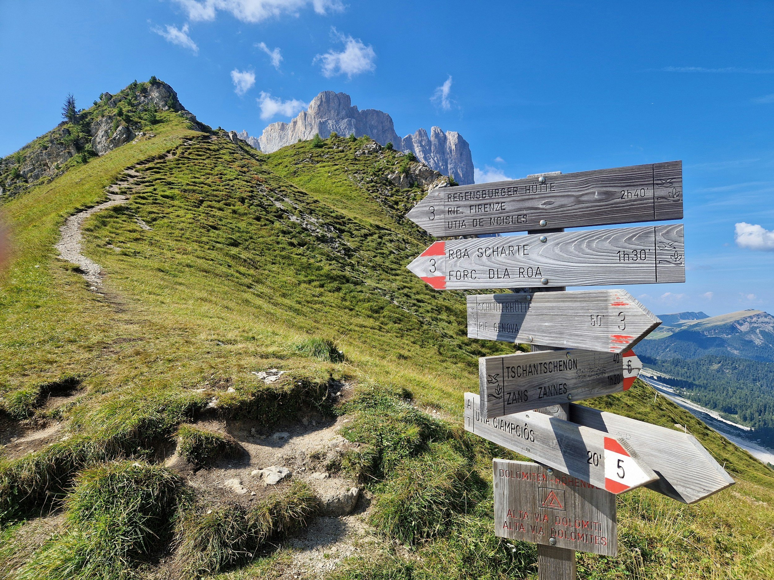 A mountain trail signpost with three directional signs, set against a backdrop of green mountainous terrain, rocky peaks, and a clear blue sky with scattered clouds.