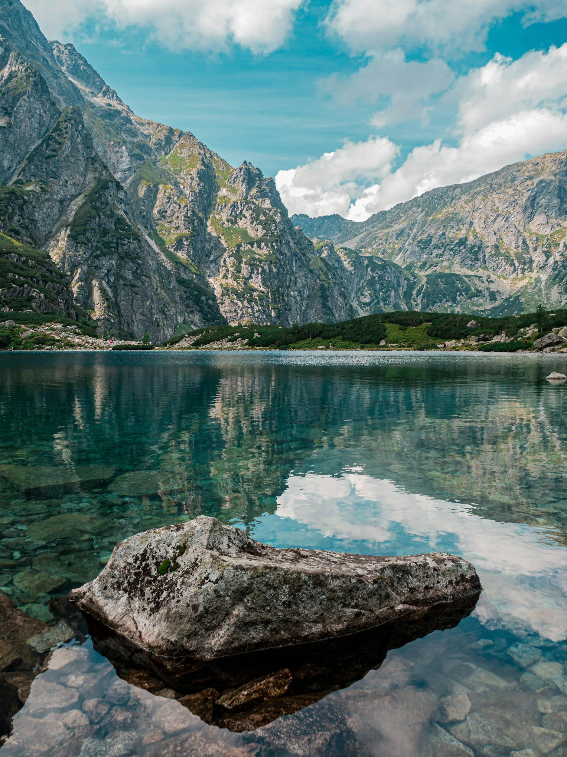 A large rock in clear water at the edge of a lake, with mountains and a blue sky with clouds in the background.