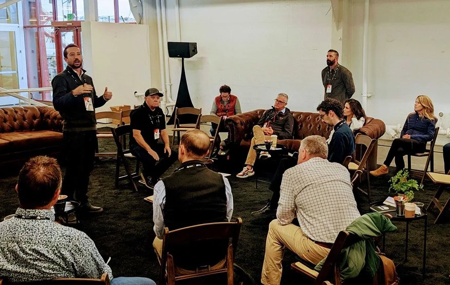 A man standing and speaking to a seated audience in an indoor conference room with a leather couch and large windows.