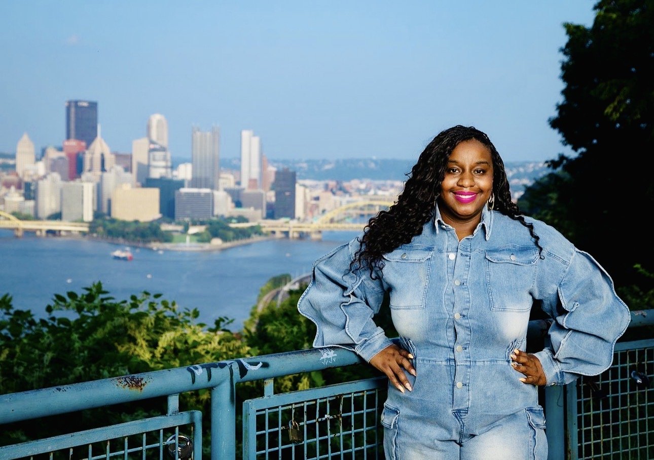 A woman standing outdoors near a blue railing with a city skyline and river in the background. She is wearing a denim jumpsuit and smiling.