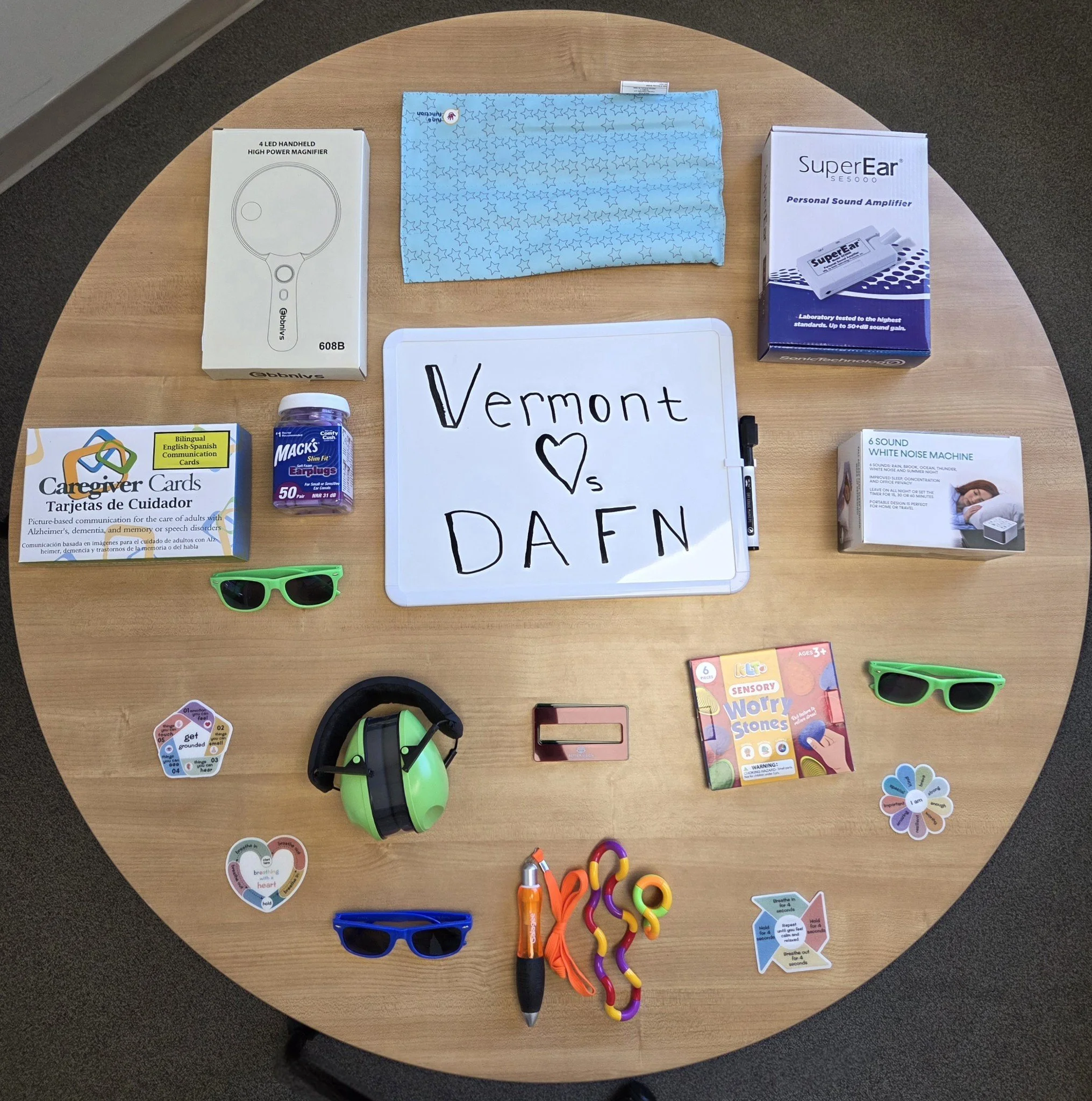 A round wooden table displaying various items including a white dry erase board with the handwritten words "Vermont ♥ s DAFN," a blue patterned cloth, a box of caregiver communication cards, a bottle of earplugs, a box of a personal sound amplifier, a box of white noise machine, two pairs of green sunglasses, a pair of blue sunglasses, a pair of black headphones, a multicolored fidget toy, colorful 'Sensory Worry Stones,' a pink stapler, a small orange and black flashlight, and some colorful rainbow-shaped and flower-shaped sensory toys.