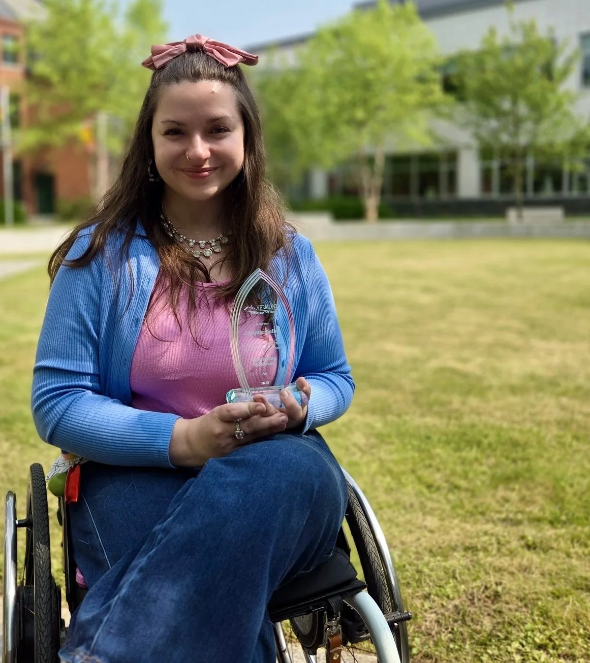 A young woman in a wheelchair outdoors on a grassy area, holding a clear glass award with the word 'Vermont' on it, smiling at the camera. She has long brown hair, wears a pink top, a blue cardigan, and a pink bow in her hair. There are trees and a building in the background.