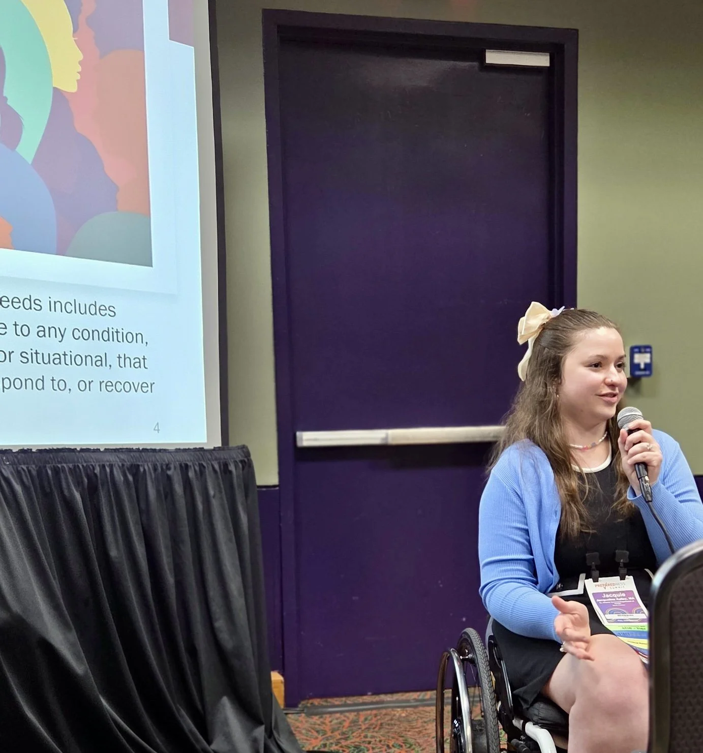 Young woman with long hair, wearing a big bow, speaking into a microphone during a presentation at a conference or seminar, sitting in a wheelchair with a attendee badge around her neck, a projected slide is visible on the screen to her left.