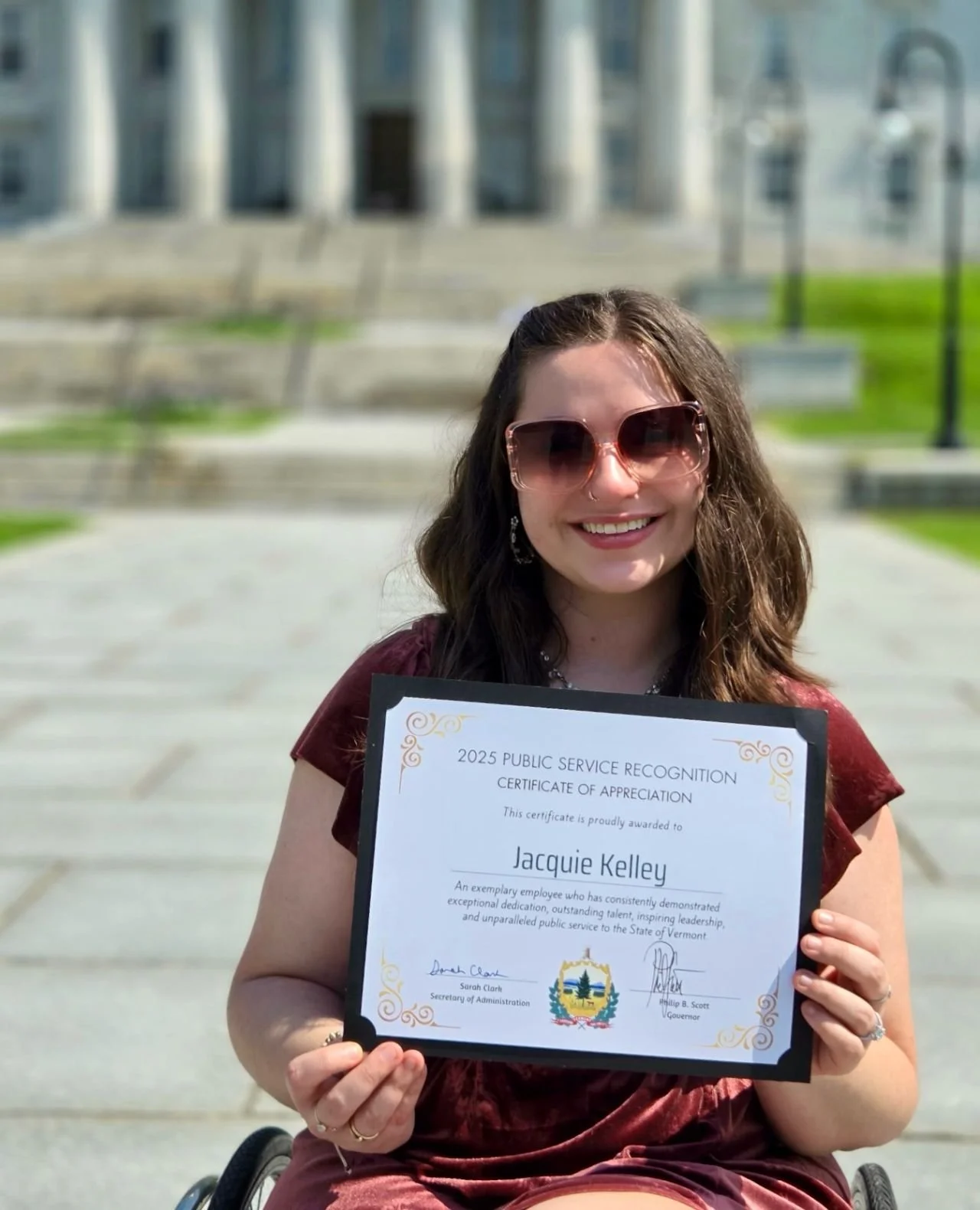 A young woman with dark wavy hair wearing sunglasses, holding a certificate of appreciation outdoors in front of a large stately building with steps, benches, and a grassy lawn.