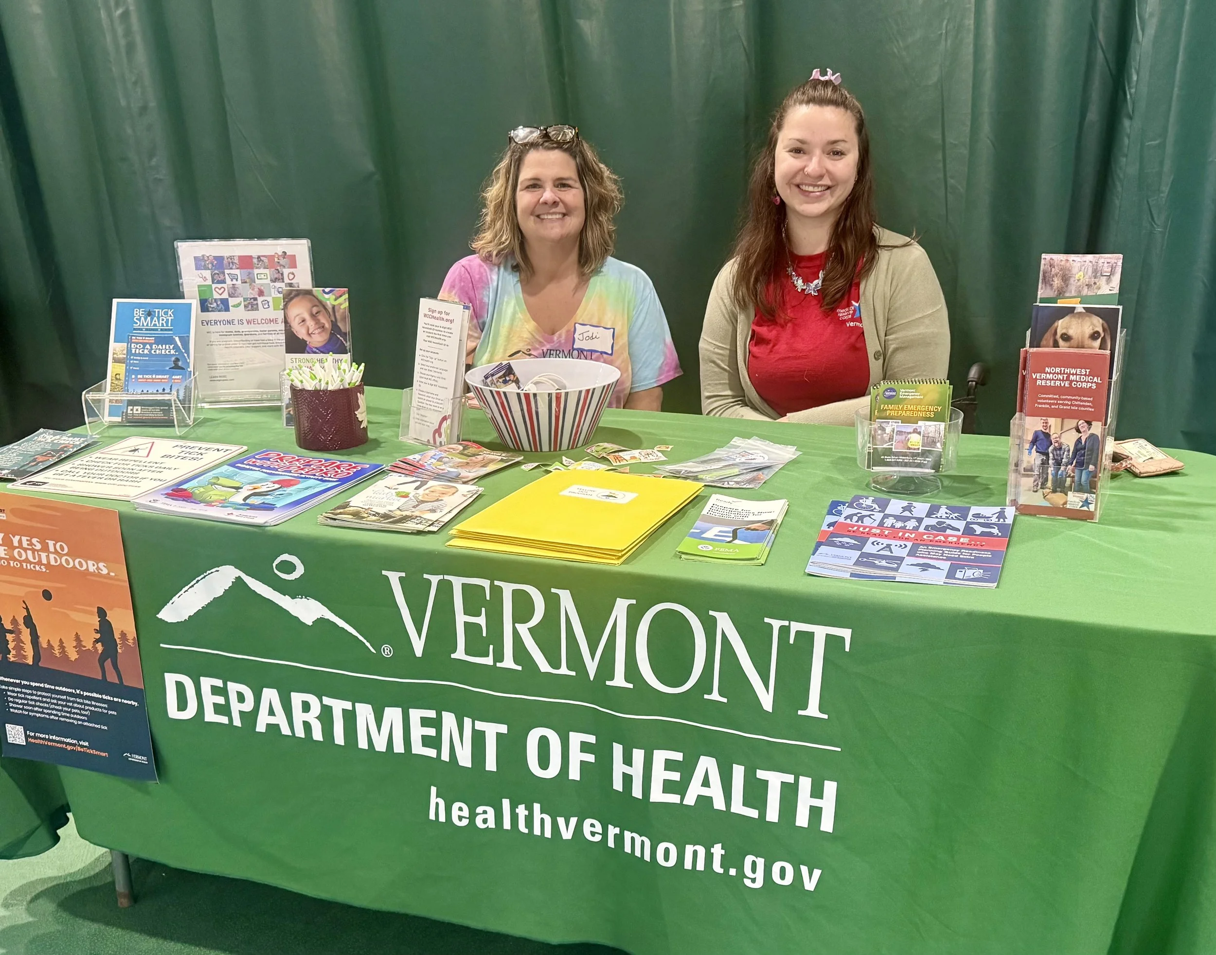 Two women sitting behind a green tablecloth with the Vermont Department of Health logo and website. The table displays pamphlets, flyers, and health information materials, with a green backdrop behind them.