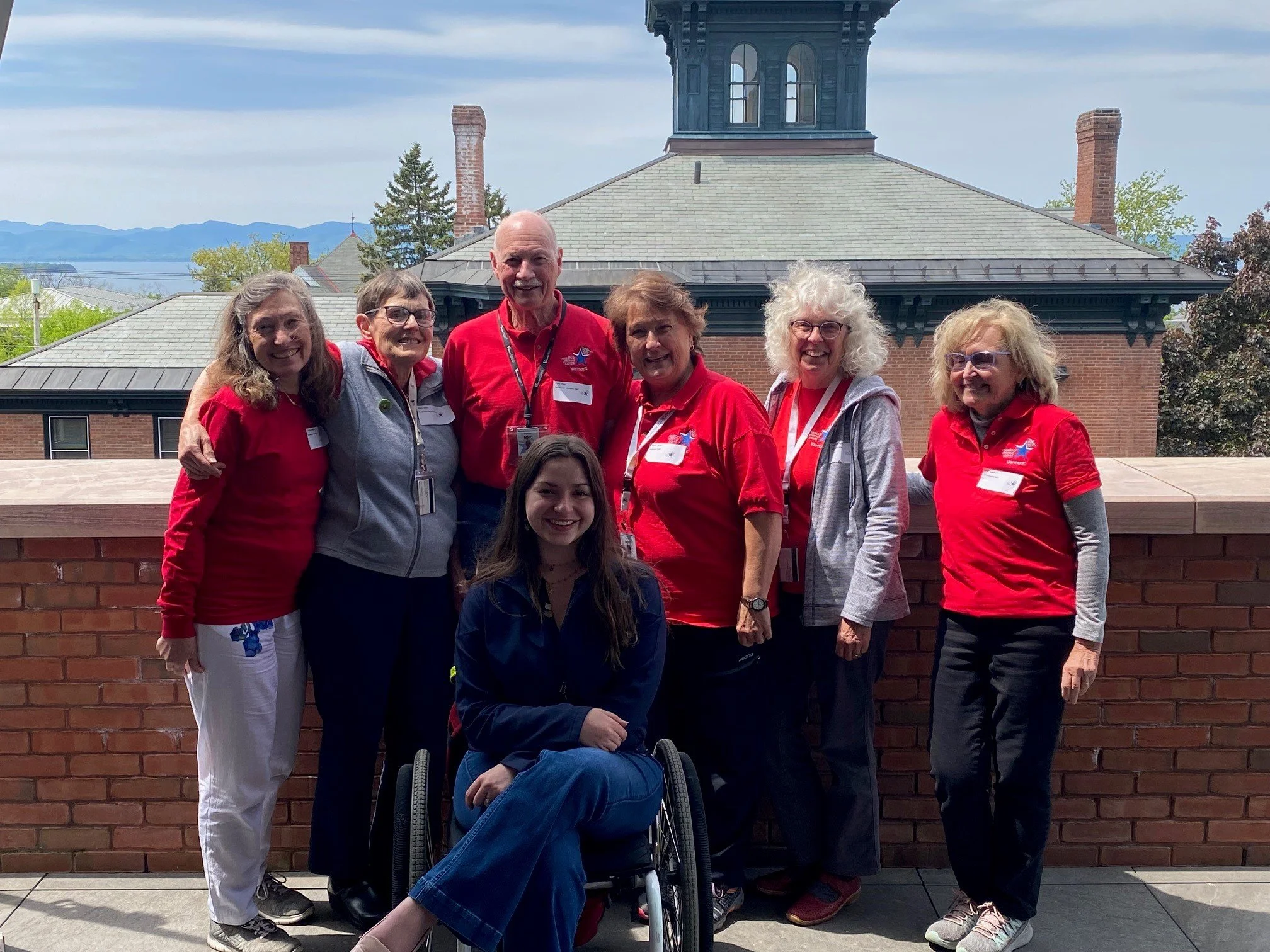 Group of eight people, mostly women, standing outdoors in front of a brick building with a tower, smiling at the camera. The group includes individuals wearing red shirts, gray jackets, and one in a wheelchair, on a balcony or rooftop with scenic mountains and trees in the background.