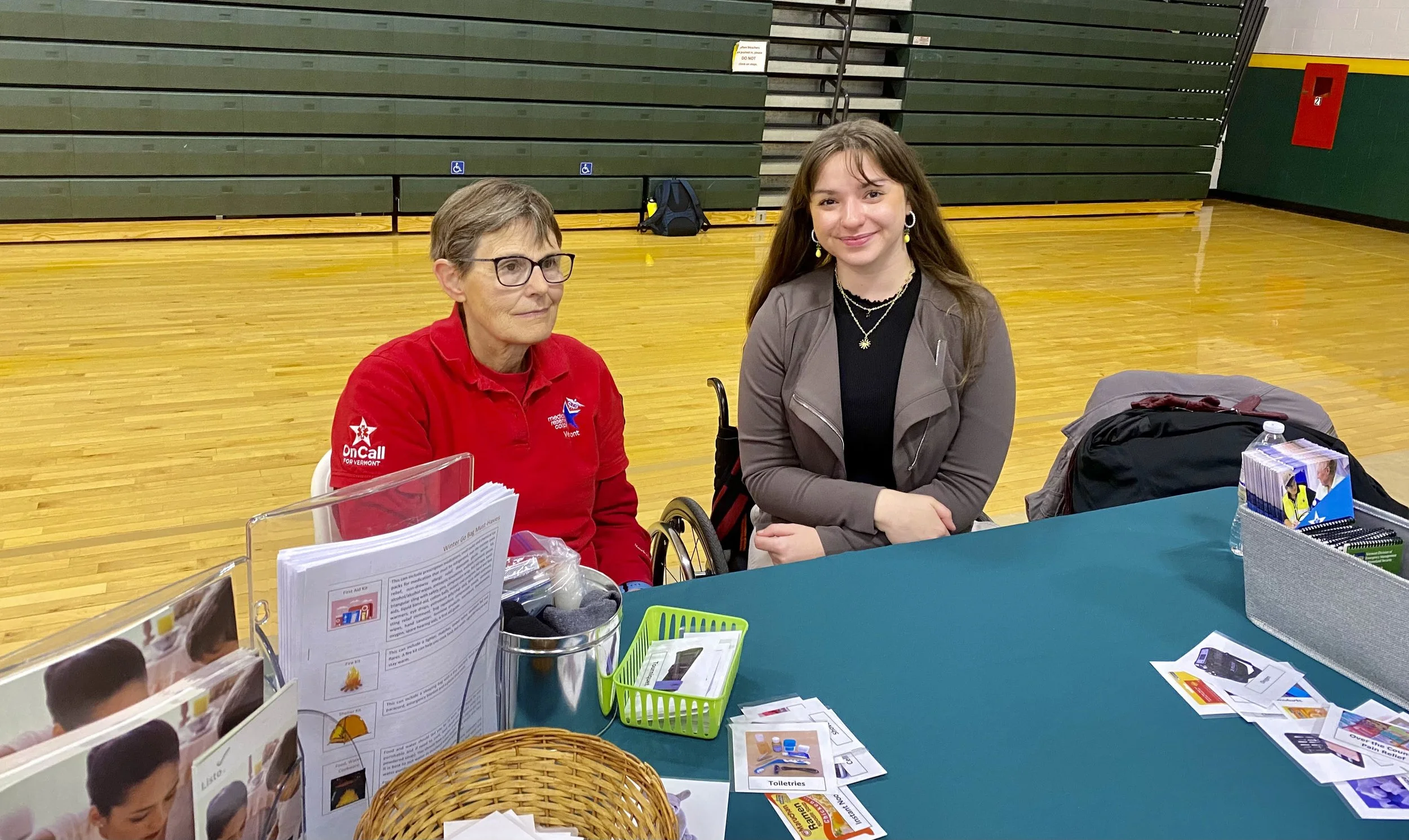 Two women sitting at a table indoors, one in a red shirt and the other in a gray jacket, with various informational materials and pamphlets on the table.