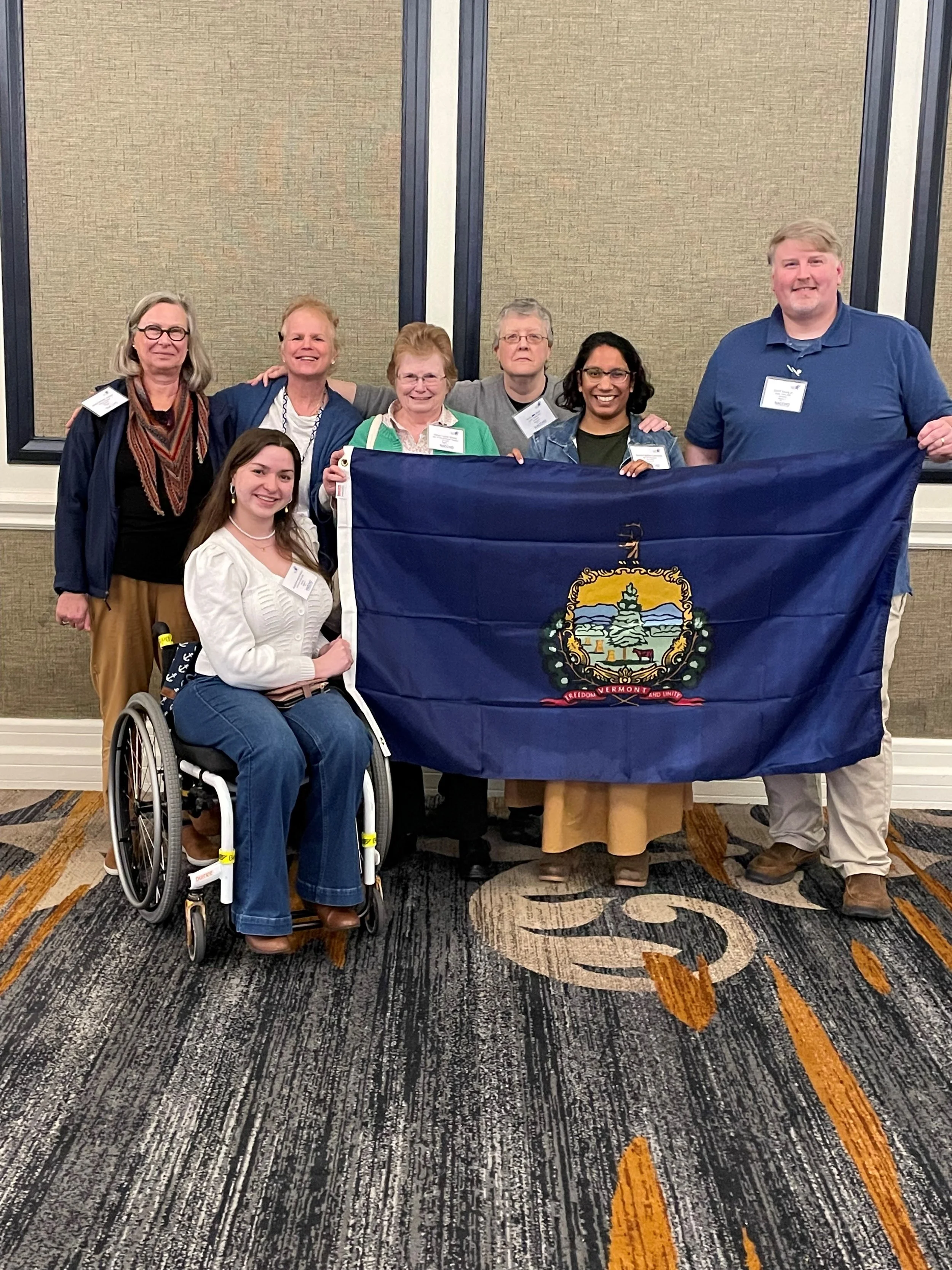 Group of seven people posing indoors behind a Vermont state flag, with a woman who is Jacquie Kelley, in a wheelchair in front, all smiling.