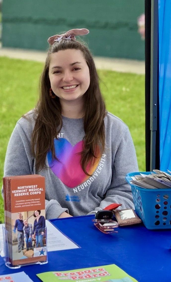 A young woman with long brown hair, smiling, sitting at a table with a green outdoor background. She has a cloth headband around her head with a small bunny ear on top. She is wearing a gray sweatshirt with a colorful heart and text. On the table, there's a brochure for the Northwest Vermont Medical Reserve Corps, a blue plastic container filled with papers, a set of keys, and a red item.