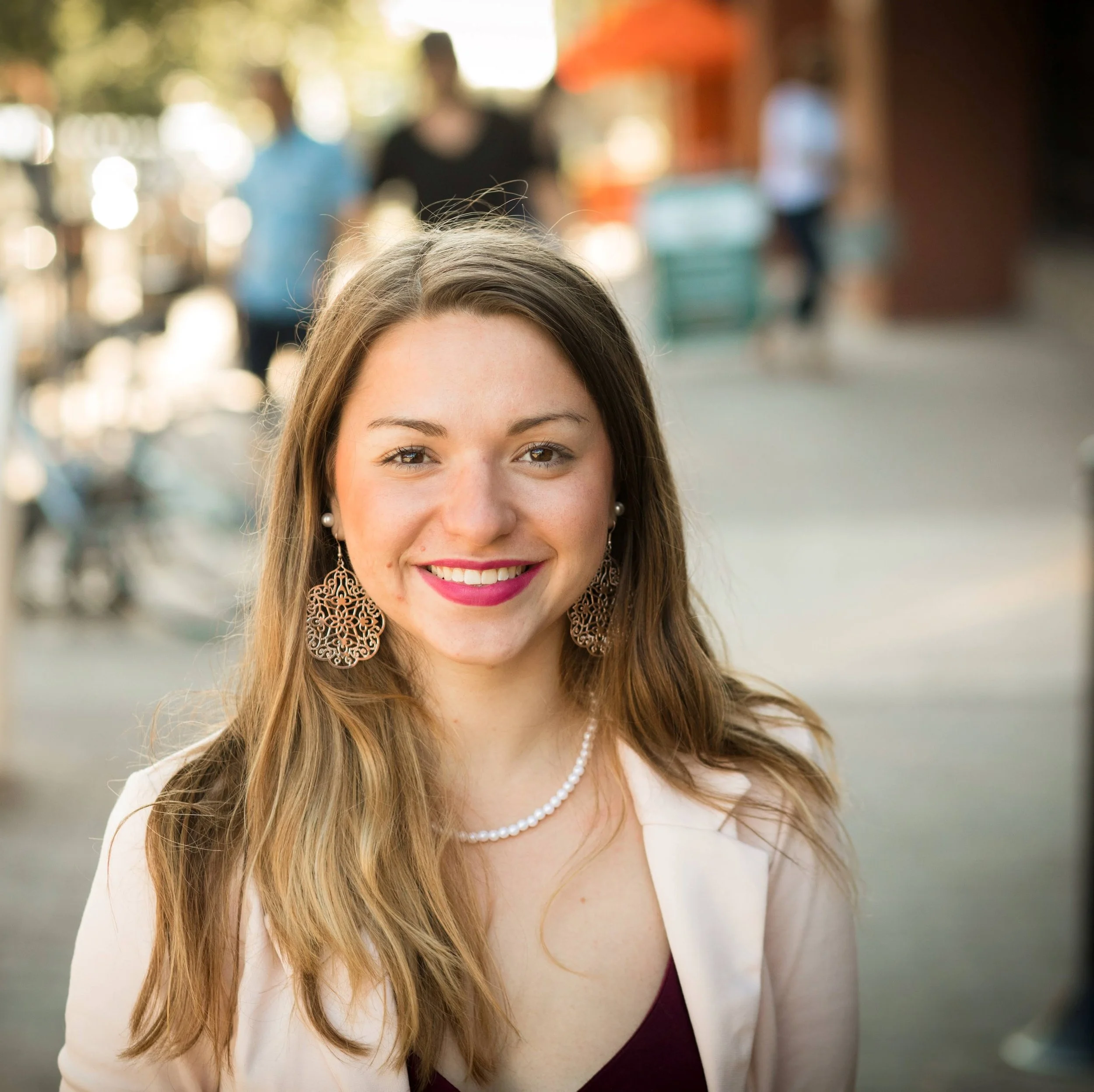 Smiling young woman which is Jacquie Kelley with long hair, wearing large earrings, a pearl necklace, and a light blazer outdoors in a sunny urban setting