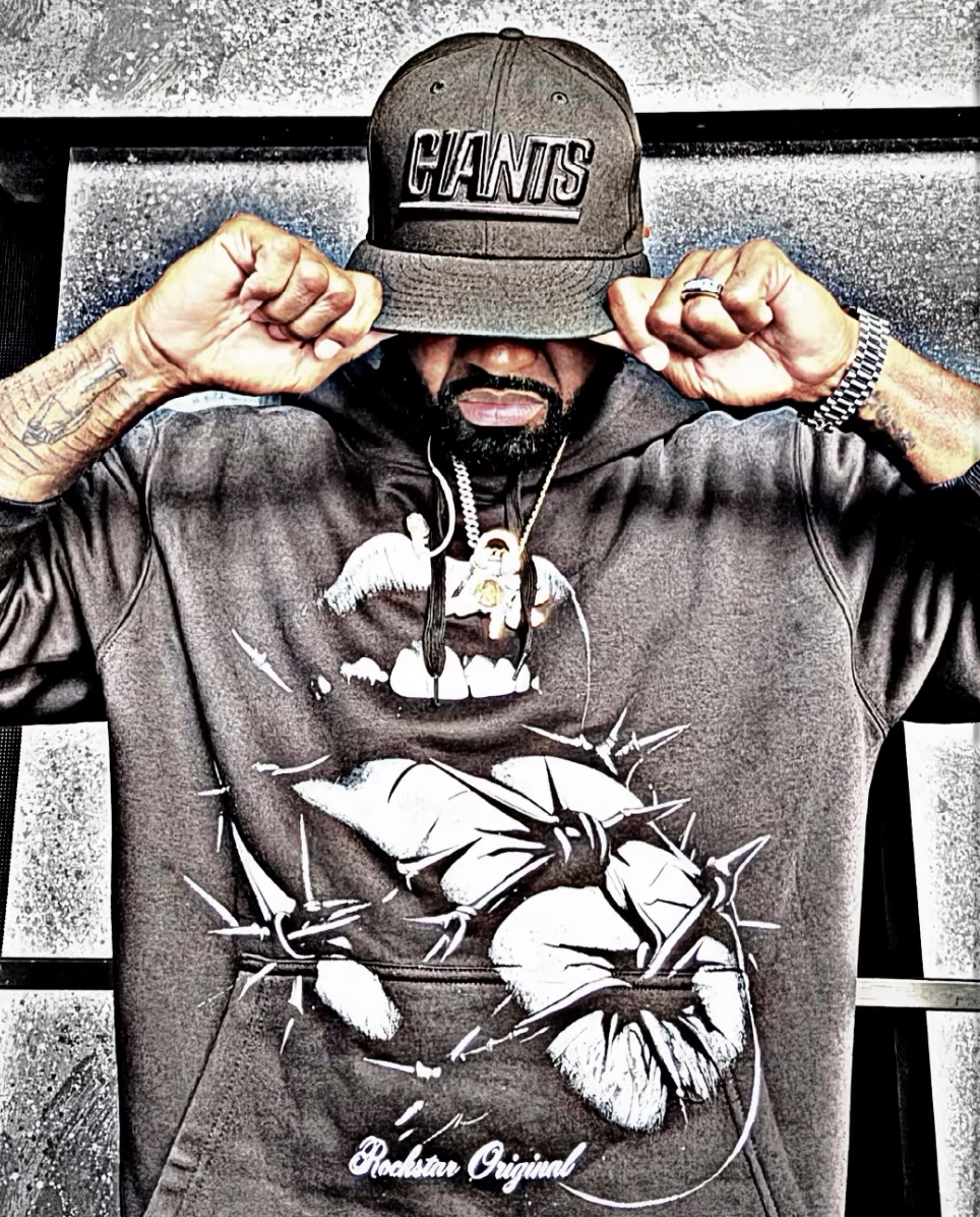 Man wearing a Giants cap and hoodie with a graphic of a face with bandages and spikes, holding the brim of his cap in a posed stance.