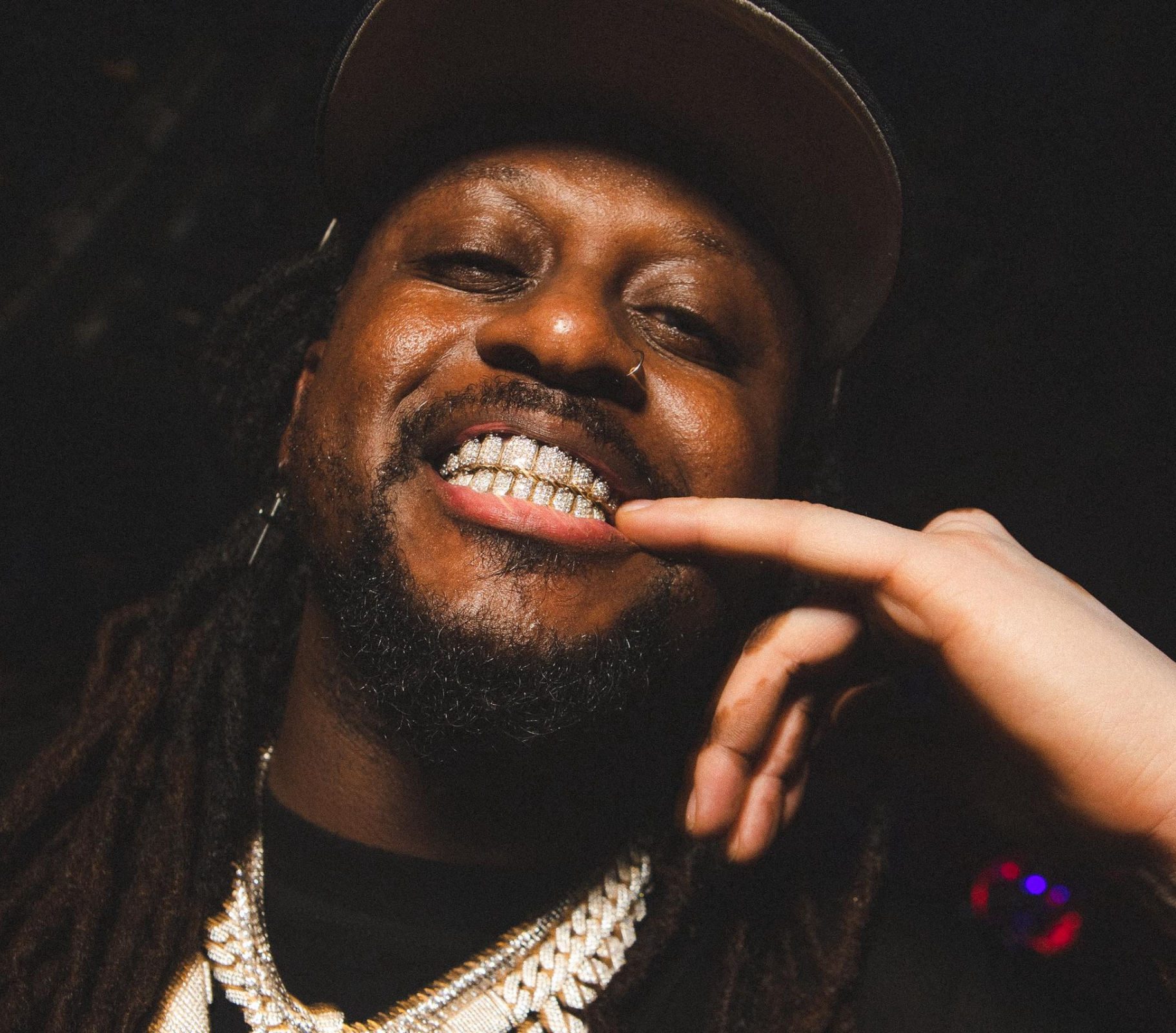 A man with long dreadlocks, gold jewelry, and a nose ring, showing off his diamond-encrusted teeth while wearing a cap.