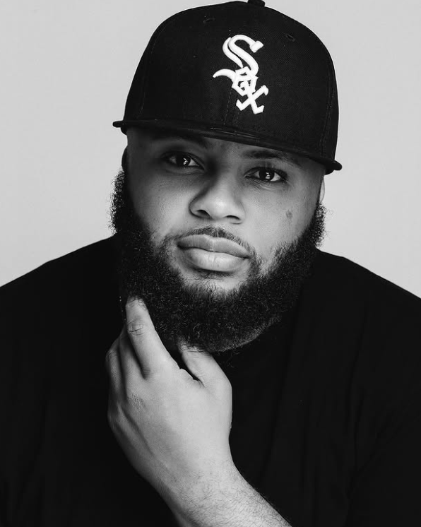 Black and white portrait of a man with a beard wearing a black baseball cap with a white logo and a black shirt, resting his chin on his left hand.