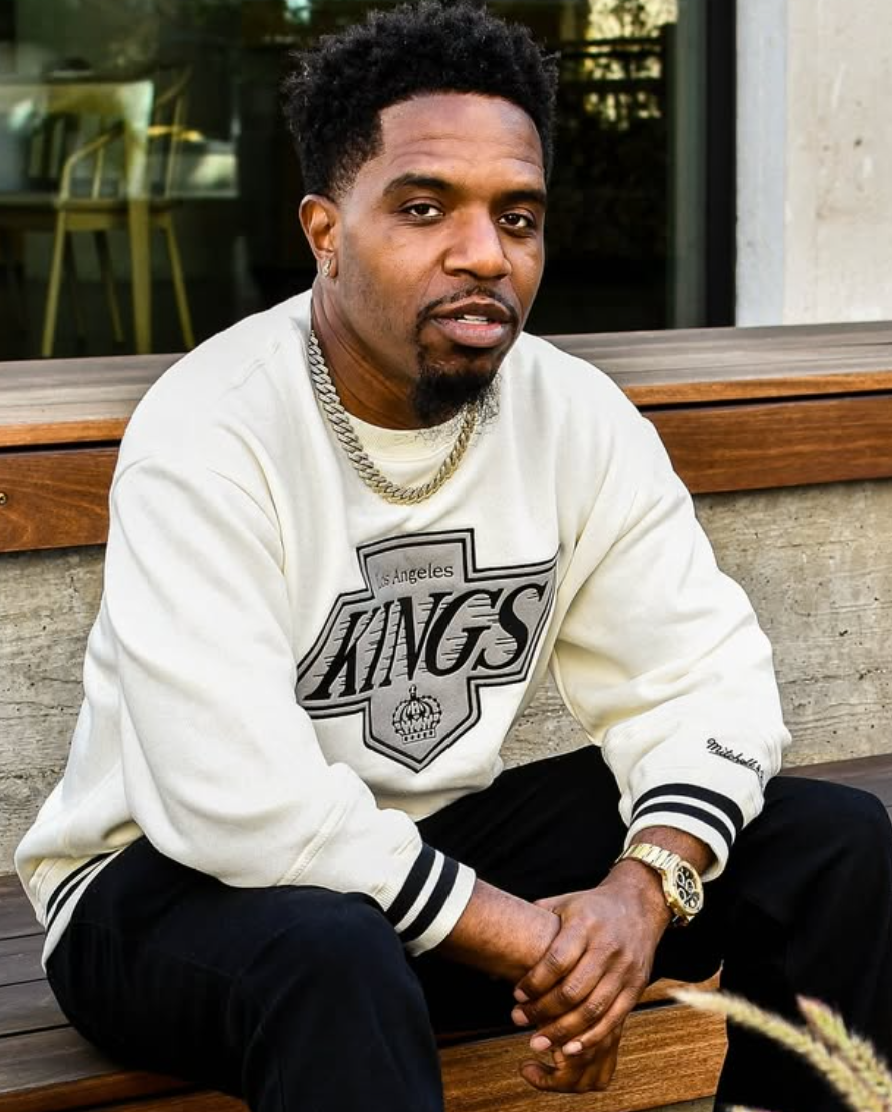 A young man sitting on a wooden bench outdoors, wearing a white Los Angeles Kings sweatshirt, black pants, gold chain necklace, and a gold watch, with short curly hair and a goatee.