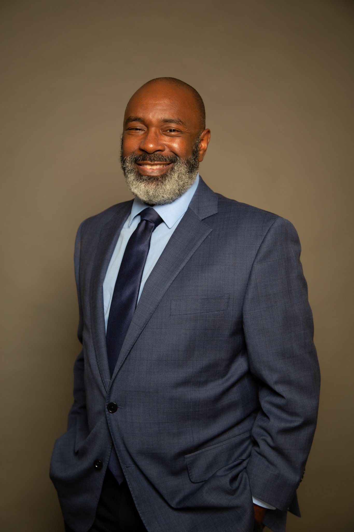 A smiling middle-aged man with a beard in a gray suit, light blue shirt, and dark blue tie posing against a plain background.