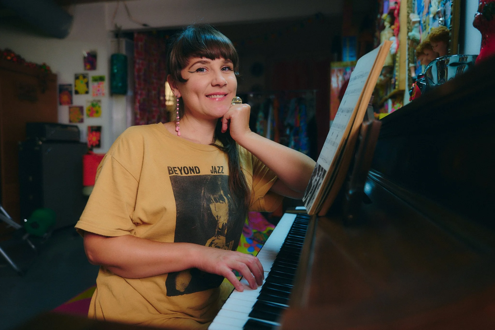 Woman with short dark hair wearing a yellow T-shirt with a jazz band print, sitting at a piano, smiling, in a colorful room with artwork and musical equipment.