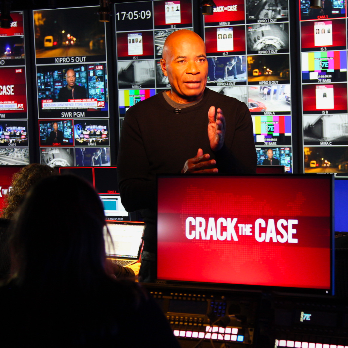 A man standing in a television studio, speaking with his hands in front of a red sign that says 'CRACK THE CASE.' Behind him are multiple screens displaying various images and footage, including news broadcasts and city scenes.