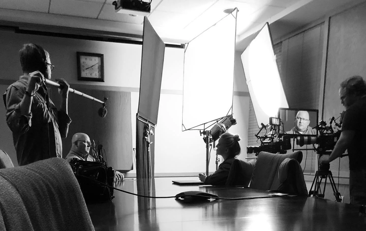 A production crew filming a woman at a conference table with a camera and lighting setup in a studio or conference room.