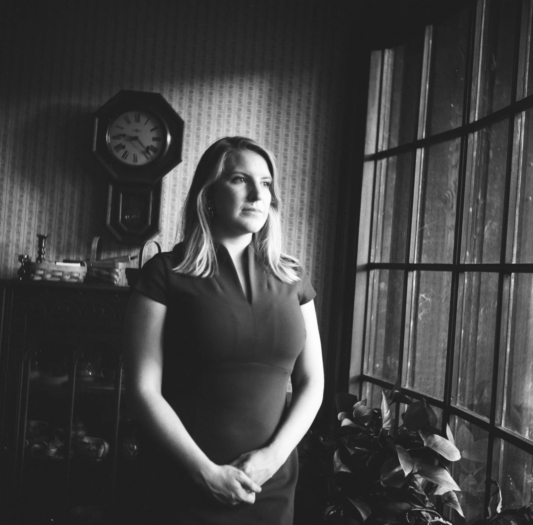 A woman with shoulder-length hair standing indoors next to a large window, looking outside. She is wearing a fitted short-sleeve dress, with her hands clasped in front of her. Behind her, there is a patterned wallpaper, a wall clock showing about 3:10, and a cabinet with various objects.