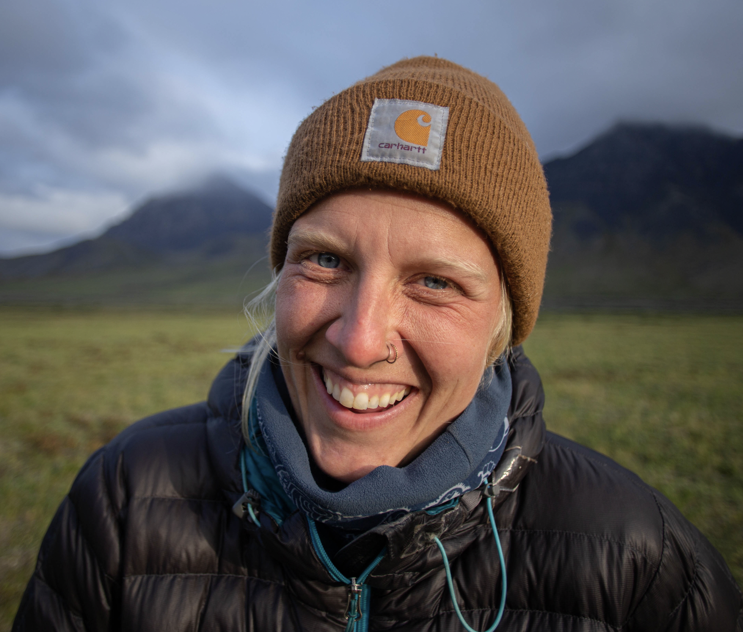 Close-up of a smiling woman with a tan Carhartt beanie, blue eyes, and a nose ring, outdoors with mountains and cloudy sky in background.