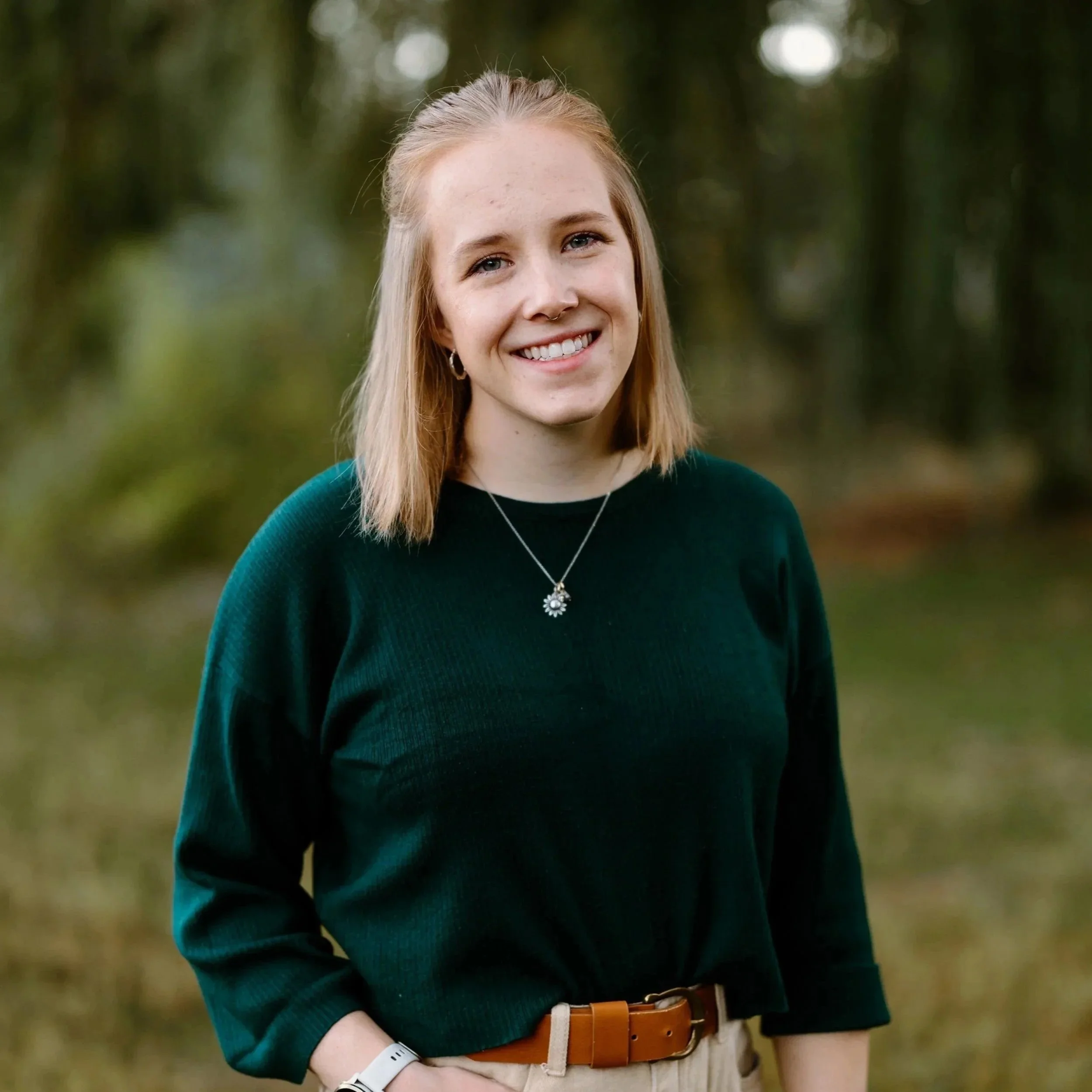 A young woman with blonde hair and a septum piercing smiling outdoors, wearing a dark green sweater, a necklace with a flower pendant, a watch, and a brown belt, standing in front of a blurred greenery background.
