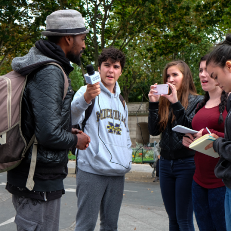 A man with a backpack and gray hat being interviewed by a group of young women and a young man, some of whom are taking notes and photos in an outdoor park setting.