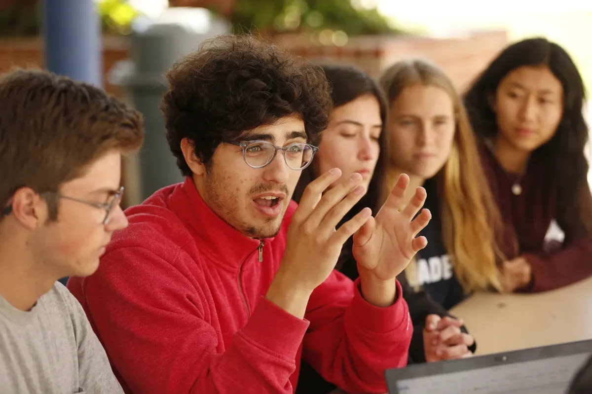 A group of five young adults sitting at a table, engaged in a discussion or meeting, with one person speaking and gesturing with their hands.