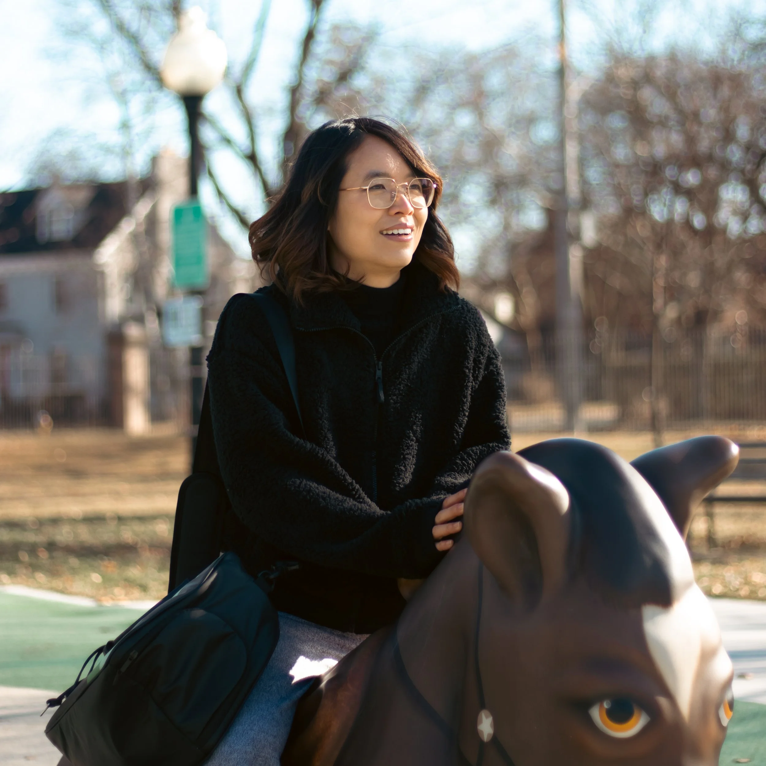 A woman with shoulder-length dark hair, glasses, and a black jacket rides a horse-shaped playground sculpture outdoors on a sunny day.