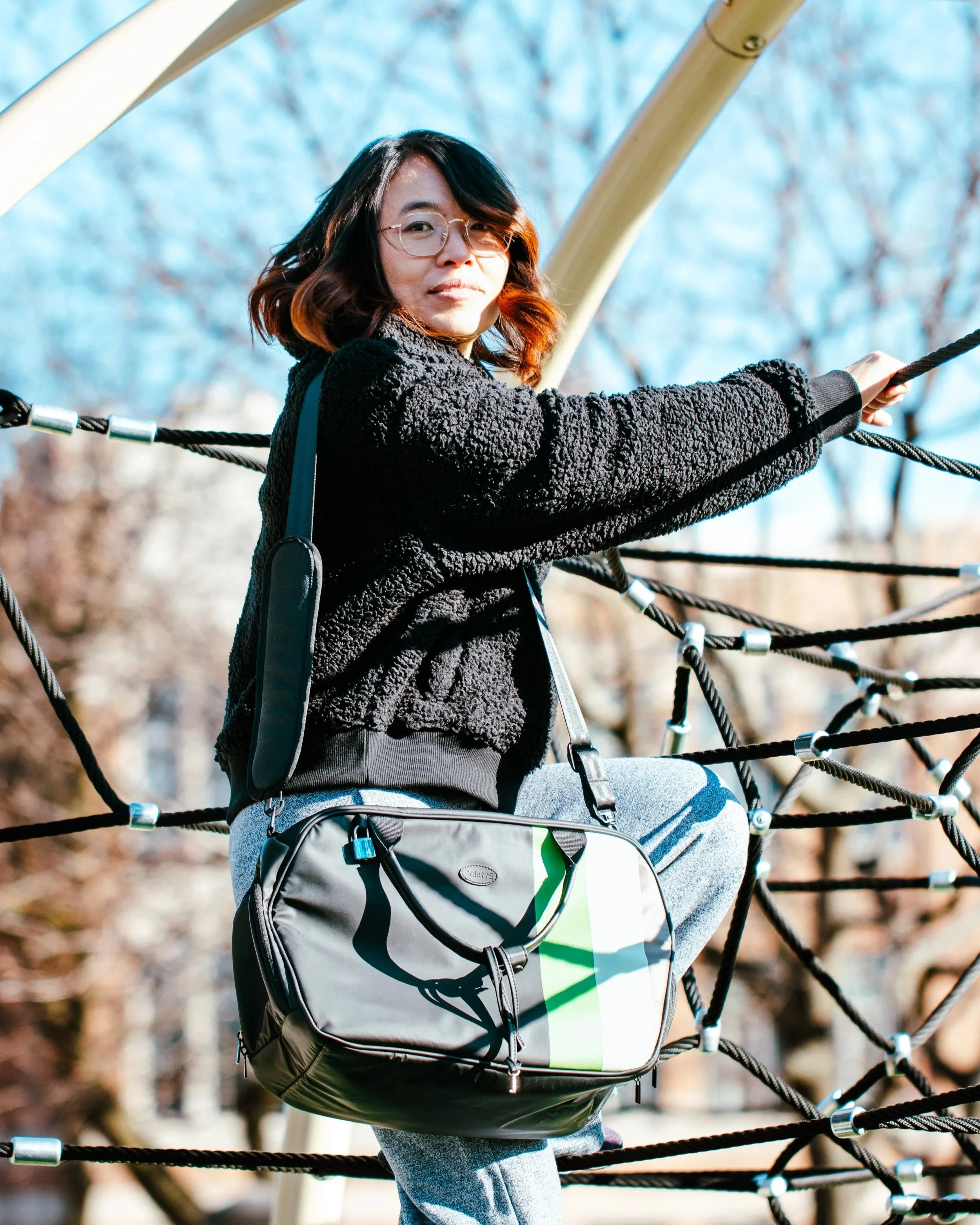 A woman with glasses and wavy hair sitting on a rope bridge outdoors, wearing a black fleece jacket and carrying a gray and green camera bag.