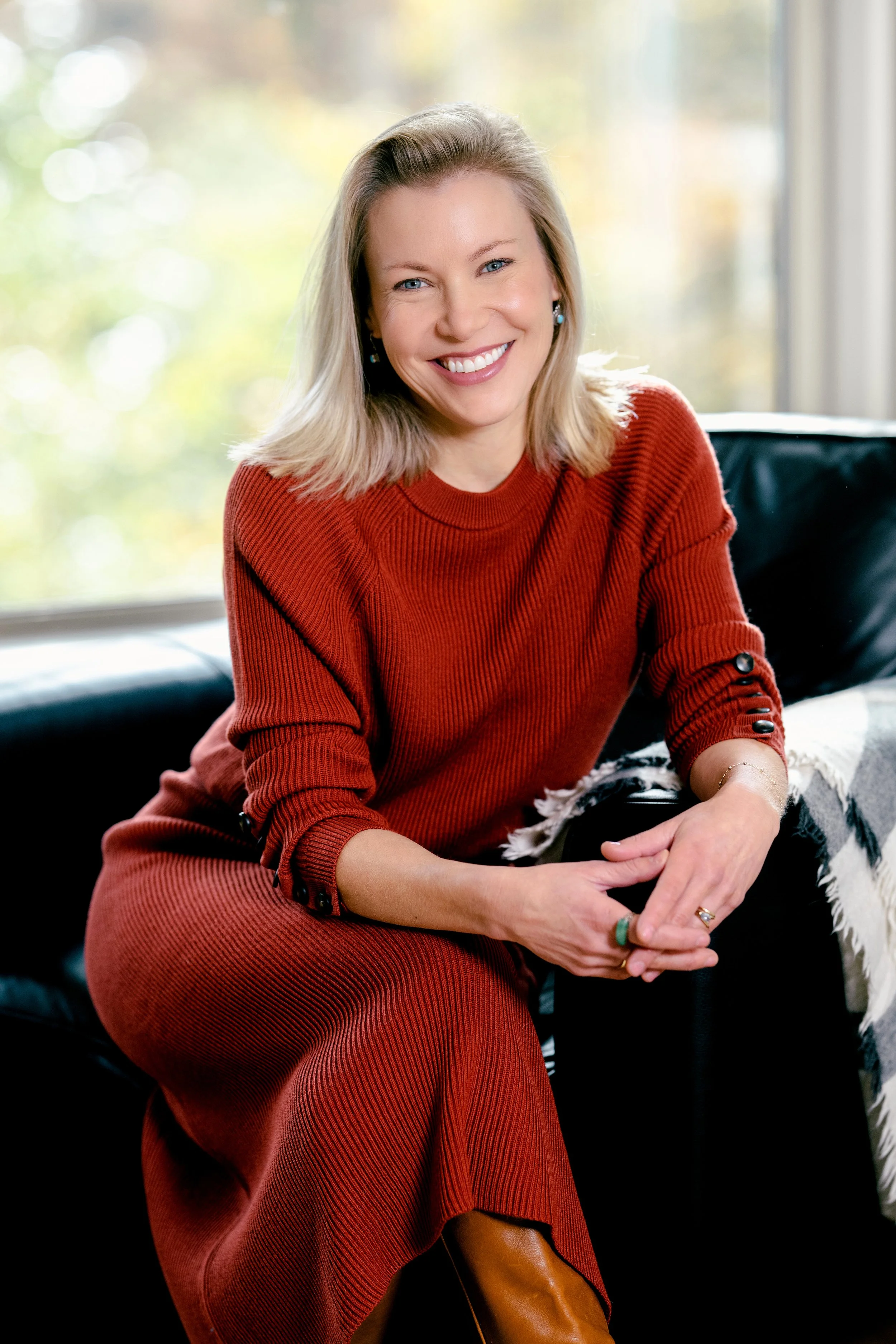 Stacey Lindsay, Author and Journalist, smiling, sitting on a black couch in front of a window with blurred autumn trees outside.