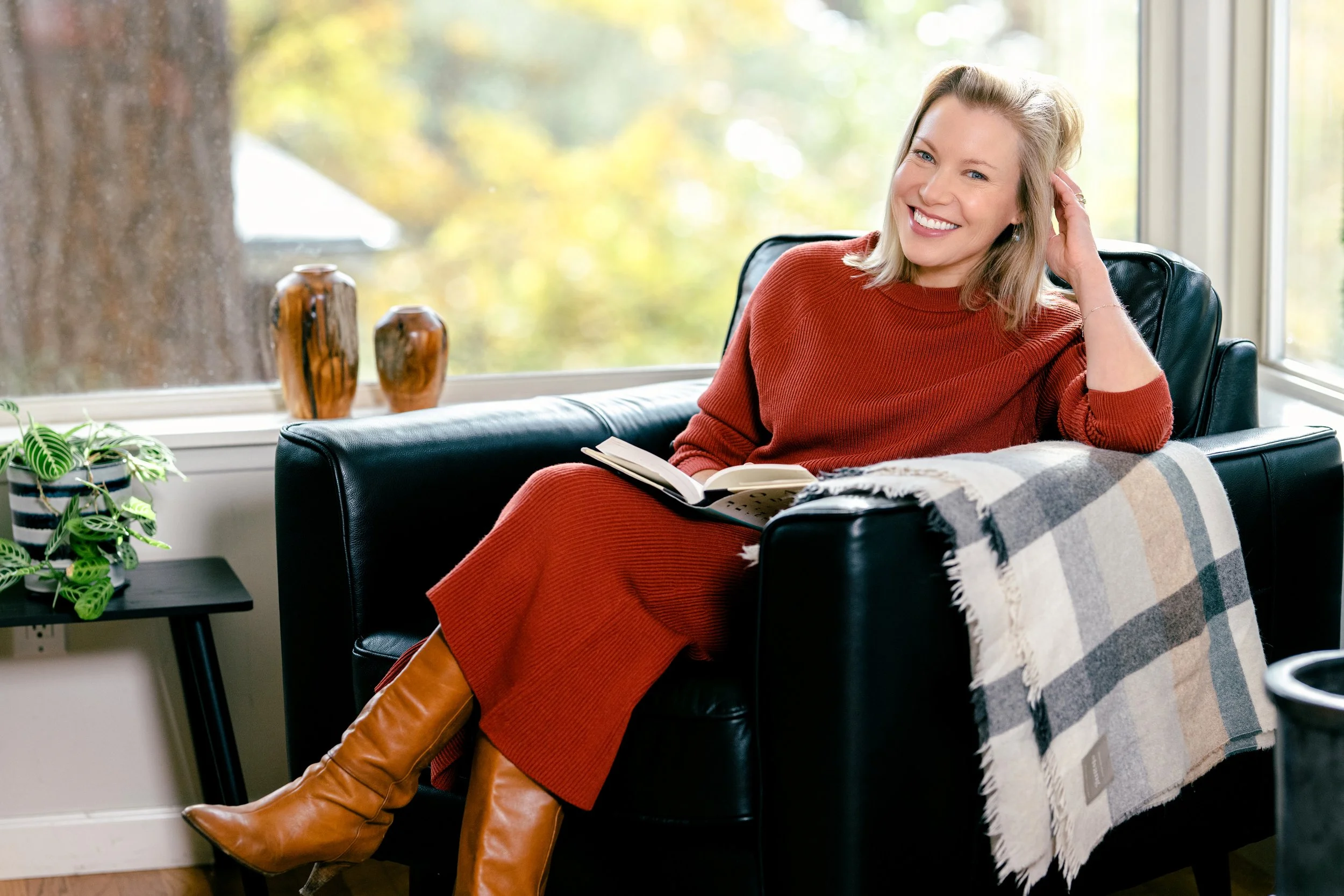 Stacey Lindsay, Author and Journalist, sitting comfortably on a black leather armchair, smiling, with a book on her lap, inside a bright room with large windows , wearing a rust-colored dress and tan boots, with a plaid blanket on the armrest.