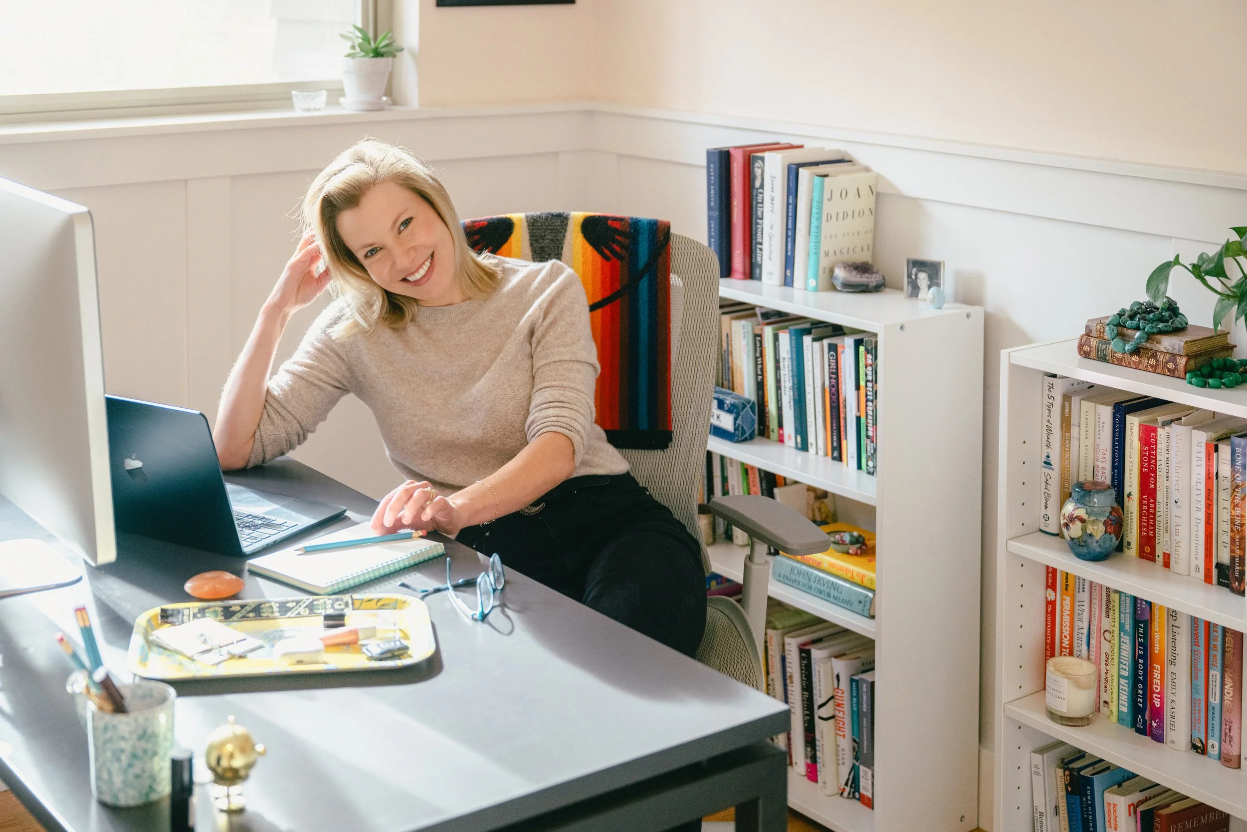 Stacey Lindsay, Author and Journalist, smiling while sitting at a desk in her home office, with a computer and bookshelves in the background.