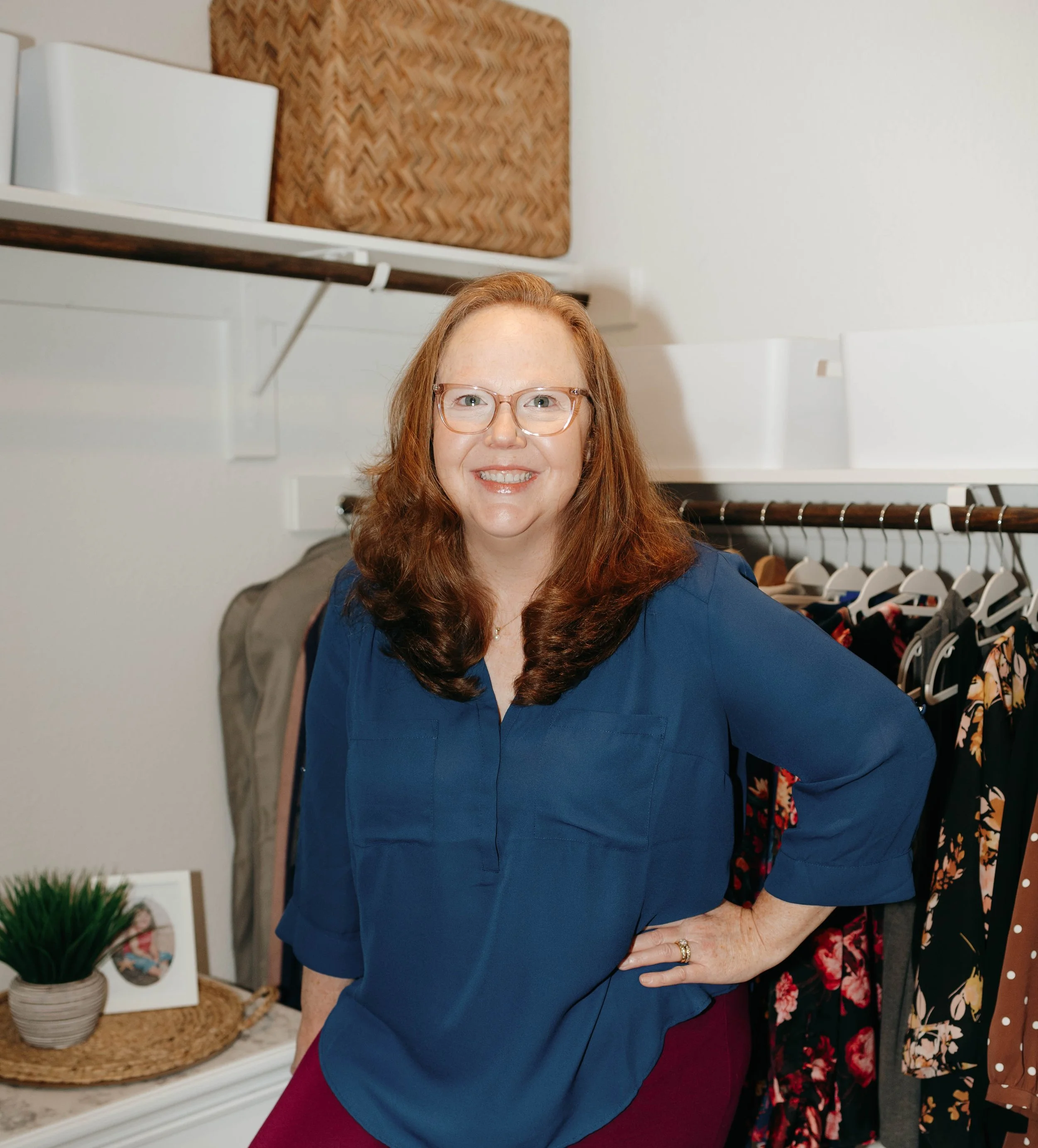 A woman with long red hair, glasses, and a blue blouse stands in a walk-in closet, smiling with her hand on her hip.