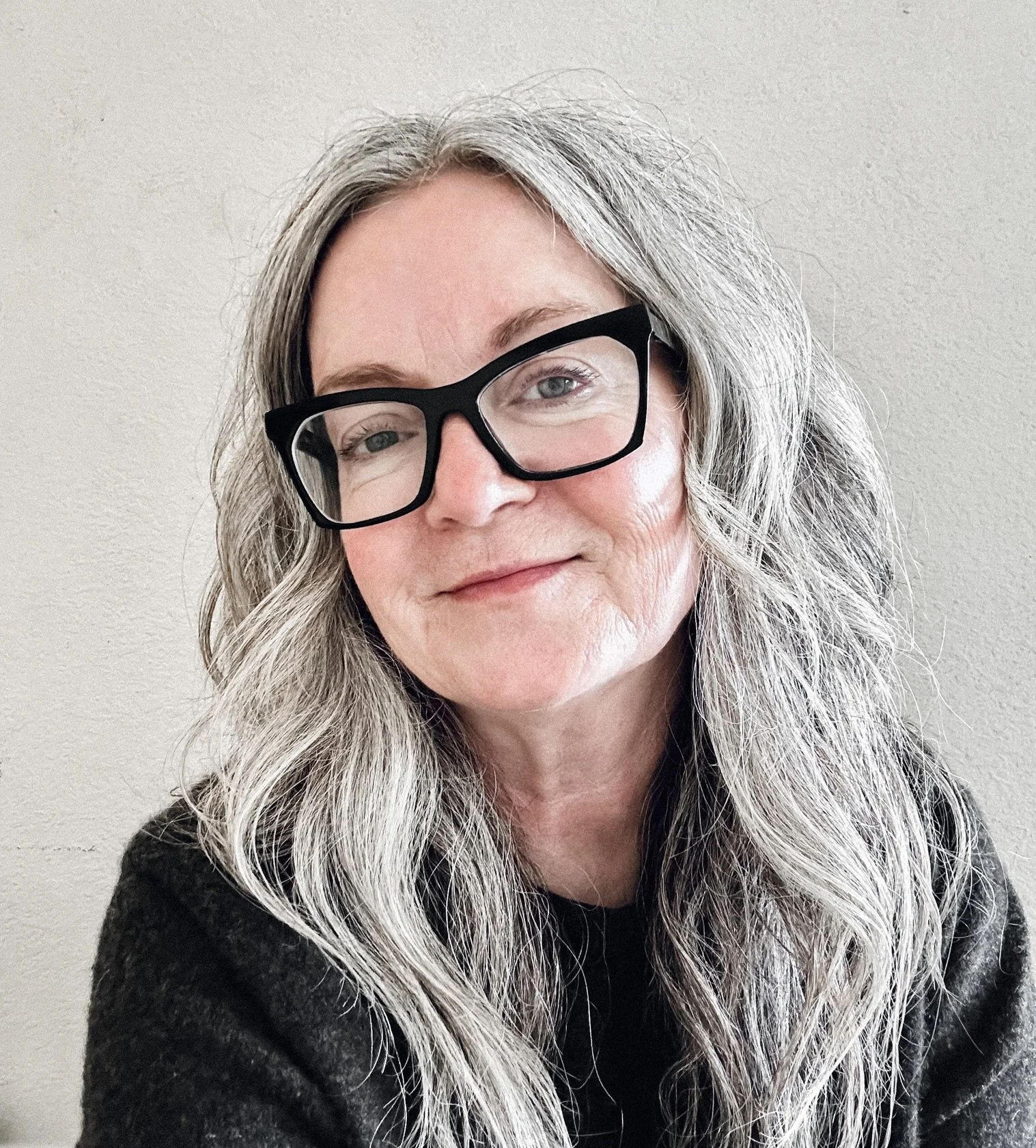 A woman with long, wavy gray hair wearing black-rimmed glasses and a black top, smiling slightly in front of a plain light-colored wall.